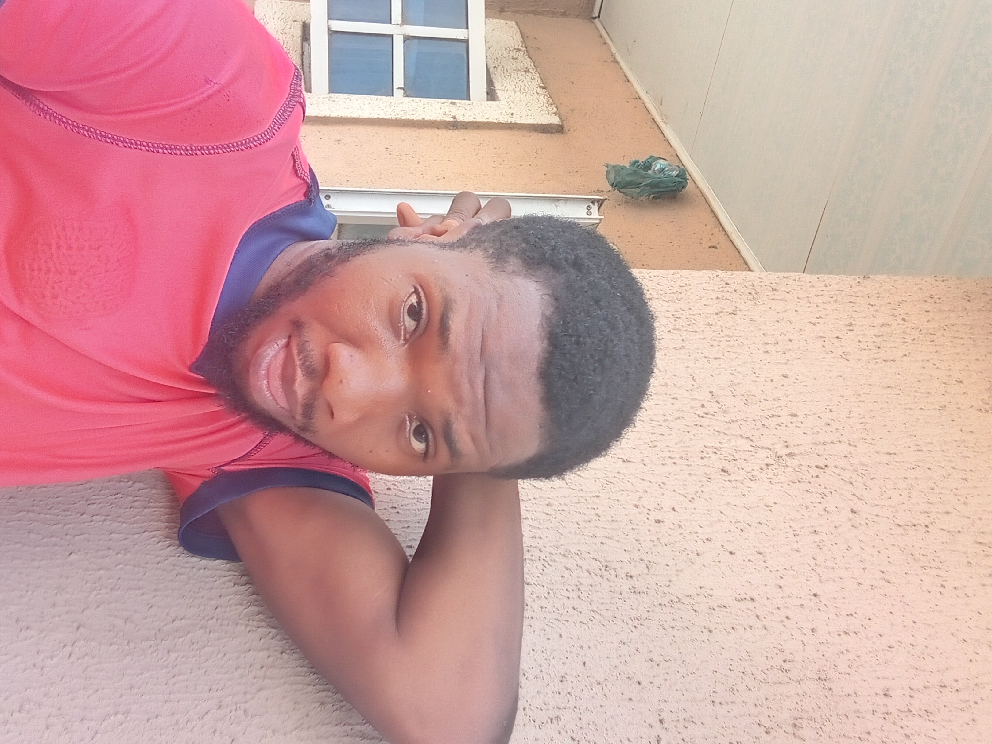 A close-up, high-angle selfie of a man outdoors in Zango-ndaji, Nigeria. He is wearing a red and blue shirt. He is lying on a light-colored surface. Above and behind him is the exterior of a building with a window featuring a white frame. A green object is attached to the wall. The surface the man is lying on is textured.