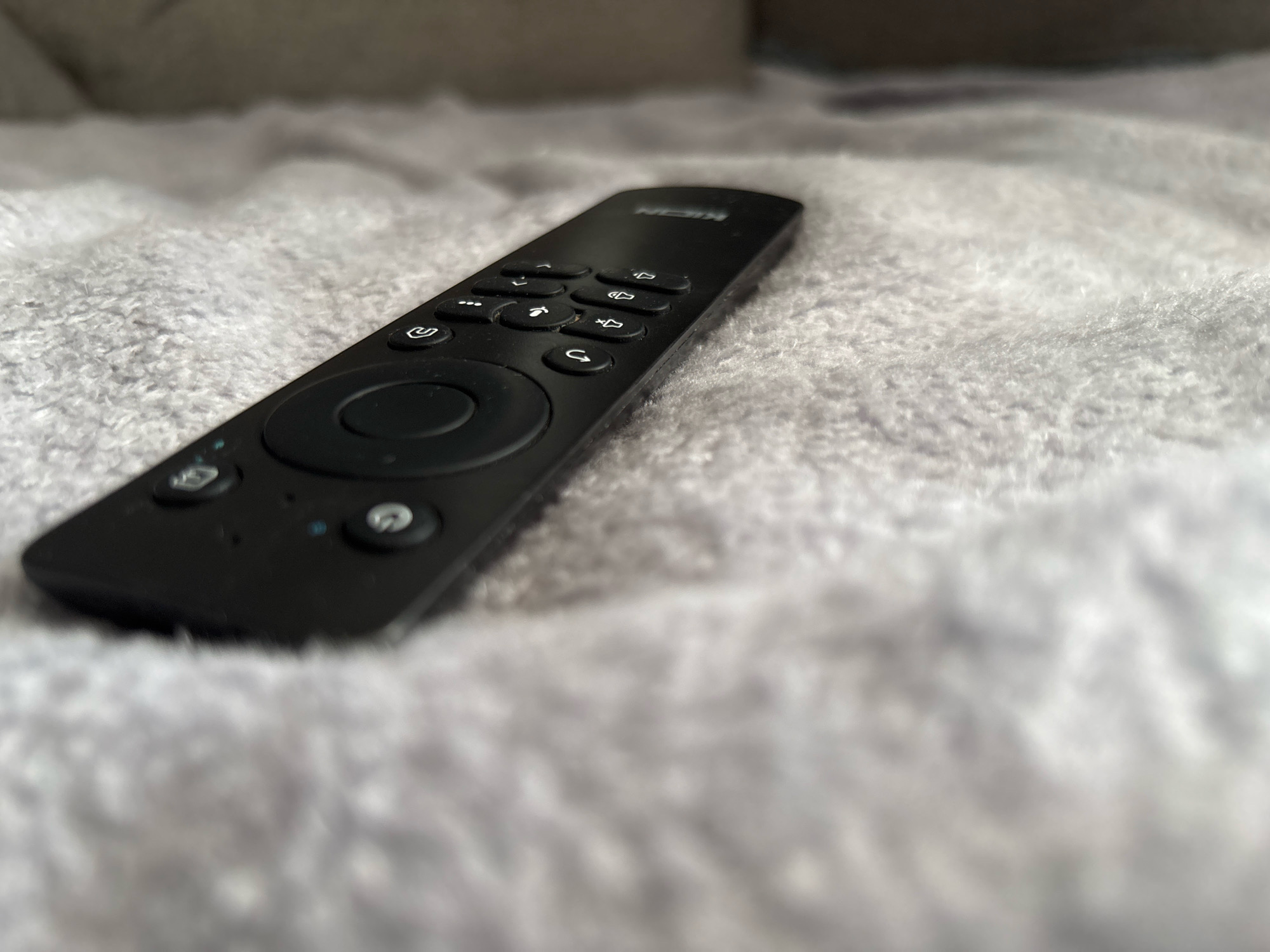 A black remote control rests on a fuzzy, light gray textured surface, possibly a blanket or rug. The remote is positioned diagonally in the frame, with its top end closer to the viewer and angled towards the right. The surface it lies on is soft and plush, with visible texture from fibers. The background is out of focus, showing a lighter, blurred area. The overall lighting is soft, with some highlights on the remote's surface. There is no indication of the city of Sinegorskiy, Russia.