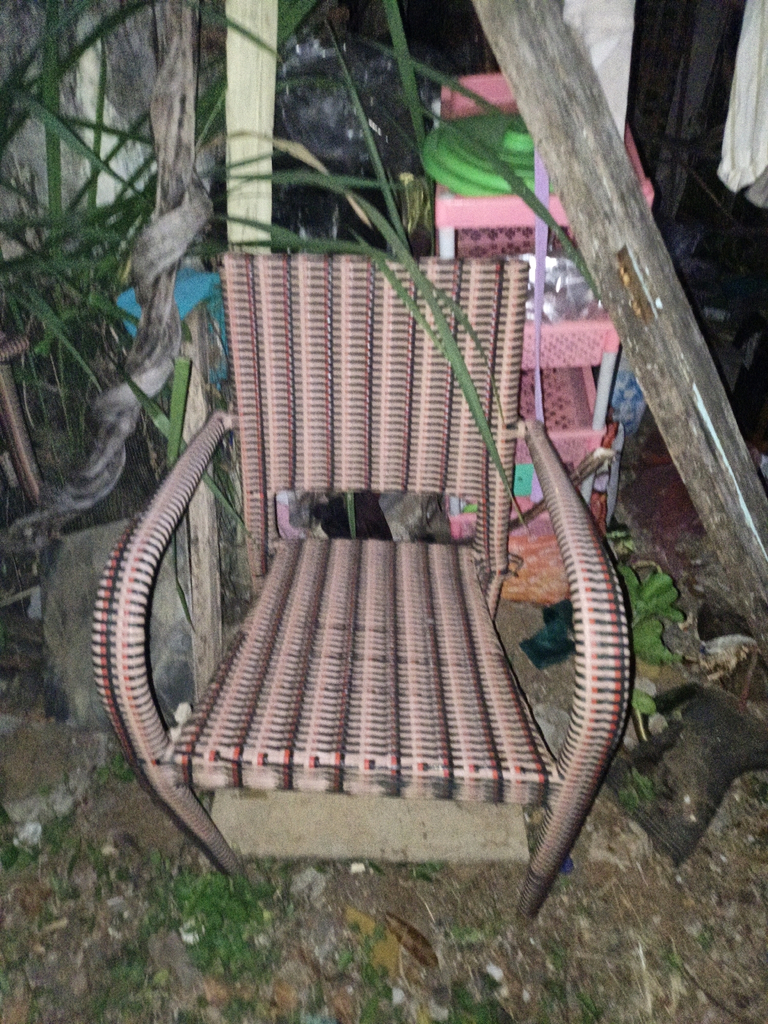 A rustic, outdoor setting in Cikal, Indonesia features a woven rattan chair with a pink and black striped pattern. The chair has curved armrests and is placed on the ground amidst overgrown vegetation and debris. In the background, there are stacked pink plastic shelves with a green item on top, a wooden beam, and some laundry hanging. The scene appears to be in disarray, suggesting a neglected or storage area. The lighting is dim, indicating it might be dusk or nighttime.