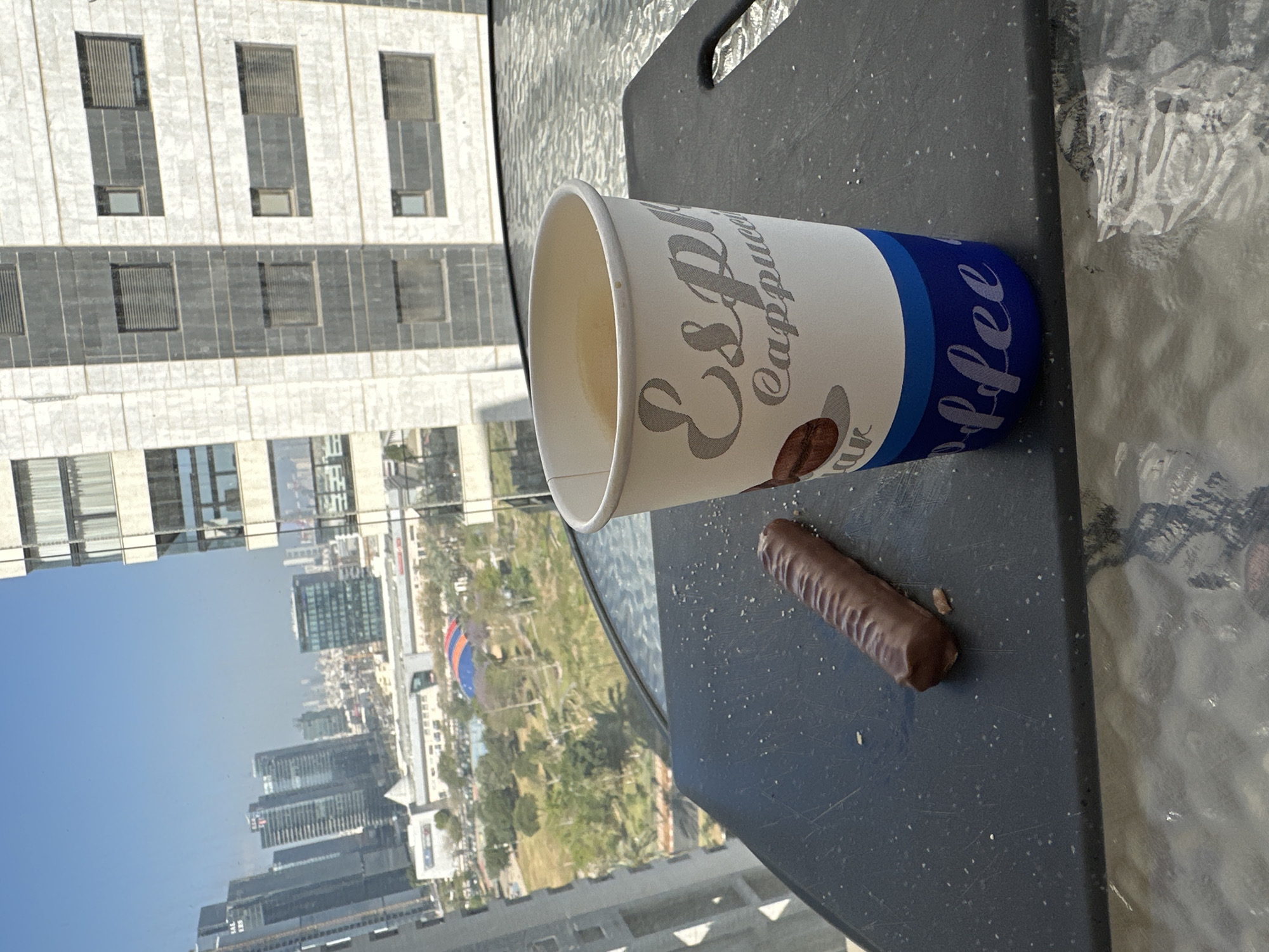 A daytime outdoor scene captured from a high vantage point, overlooking an urban landscape, likely in Petah Tikva, Israel. In the foreground, a white disposable coffee cup and a ribbed chocolate bar rest on a dark, possibly plastic, serving tray or surface. The tray is placed on a glass table, which reflects and distorts the city view below.
The coffee cup features a graphic of a coffee bean and prominent text. "ESPRESSO" is written in a stylized, slightly distressed font, with "Cappuccino" in a flowing script below it. A blue band encircles the lower part of the cup, displaying the word "Coffee" in white, cursive-like lettering. The cup appears to contain a light-colored beverage. The chocolate bar is dark brown and has a series of longitudinal ridges.
The background reveals a clear blue sky above a sprawling city. Numerous multi-story buildings with varied architectural styles and window patterns are visible. One prominent building in the mid-ground features a white facade with dark horizontal bands and rectangular windows. Further in the distance, more buildings and a green area with trees can be seen. A large, rounded structure with blue and orange sections is noticeable in the middle distance. The scene suggests a sunny day with good visibility.