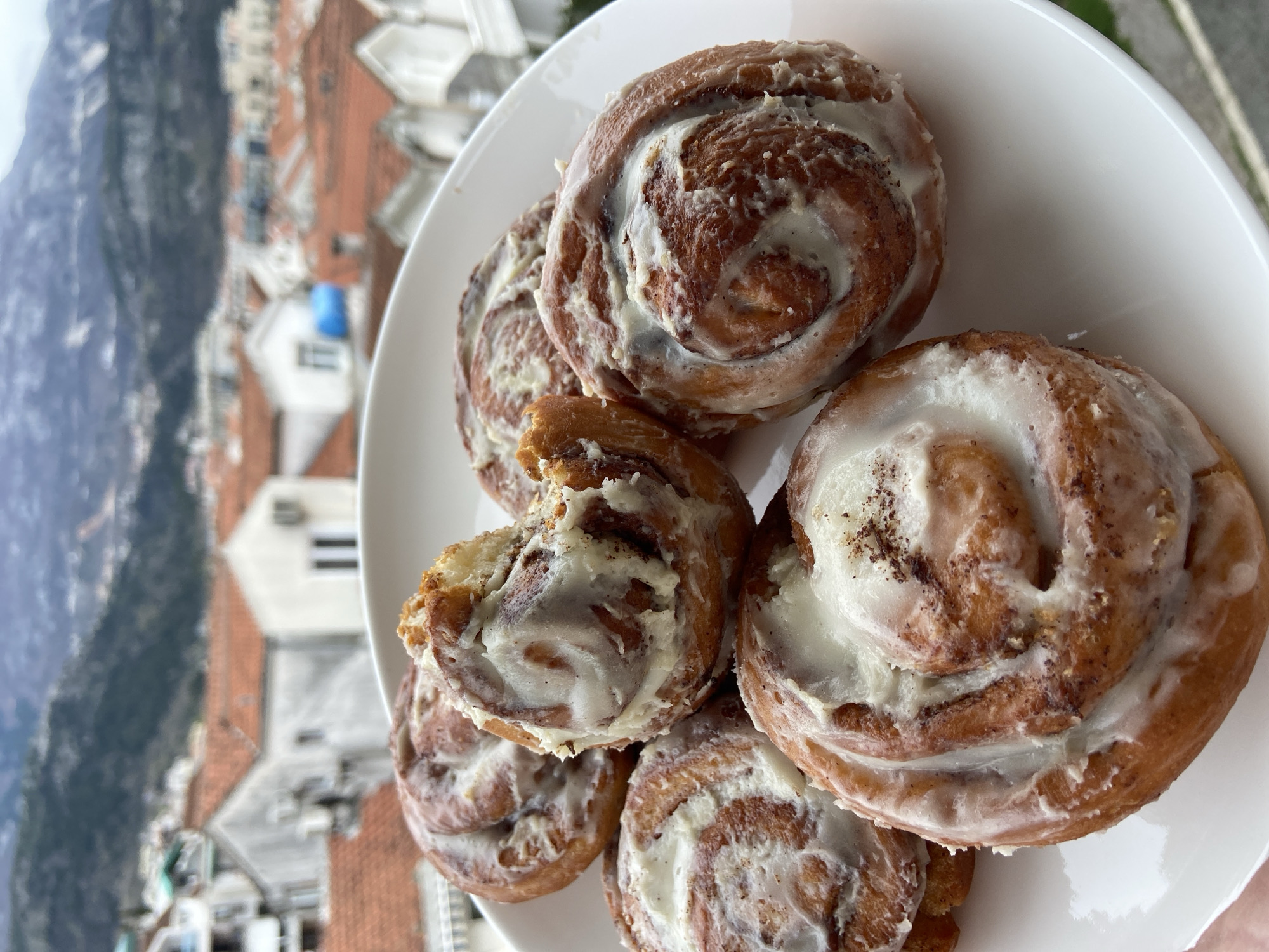 A top-down, close-up shot features a white plate piled with five cinnamon rolls, one with a bite taken out, topped with white icing. The background is blurred, showing red-tiled rooftops and a mountain, suggesting the location is Budva, Montenegro. The focus is entirely on the pastry arrangement on the plate.