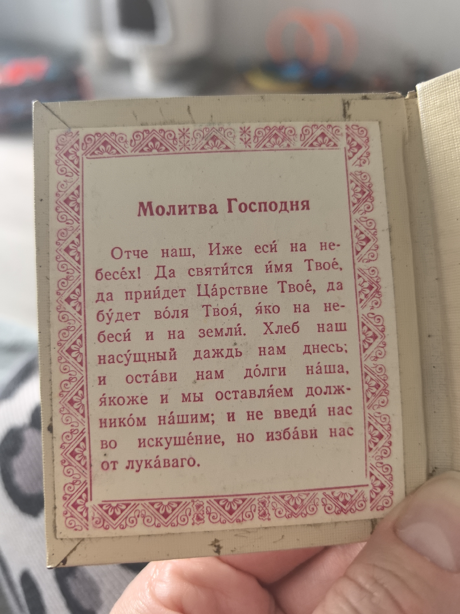 The image shows a close-up view of a page from a small book or booklet. The page has a cream-colored background with a pink decorative border. Centered on the page is text in the Russian language. The title, written in a slightly larger and bolder font, reads "Молитва Господня," which translates to "The Lord's Prayer". Below the title is the text of the prayer itself. The prayer is written in pre-1918 orthography based on the ending of "бесех!" which uses the now-defunct "ѣ" or "yat" letter. Given the text content, it is likely from an Orthodox Christian prayer book. The page appears to be held by a person, whose thumb and part of their hand are visible in the foreground. The background is blurry and out of focus, but it includes a hint of furniture and other objects, suggesting an indoor setting. There is no specific evidence within the image to suggest a location in Perm, Russia.