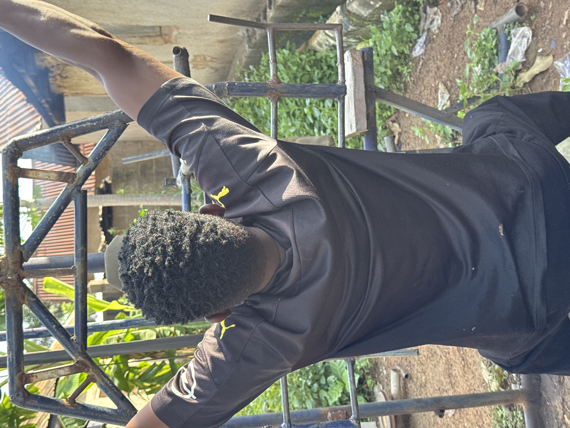 A young Black male, viewed from the back, is using a homemade weightlifting apparatus. He has short, dark, curly hair and is wearing a dark t-shirt with yellow accents and dark pants. His arms are raised above his head, gripping the metal bars of the equipment. The apparatus appears to be constructed from various metal tubes and bars, with some rust visible. He is positioned outdoors, in what appears to be a rural or semi-rural setting in Akwanga, Nigeria. The ground is dirt, with some scattered debris and vegetation. In the background, there are buildings with corrugated metal roofs and more greenery. The lighting suggests it is daytime, with sunlight casting shadows.