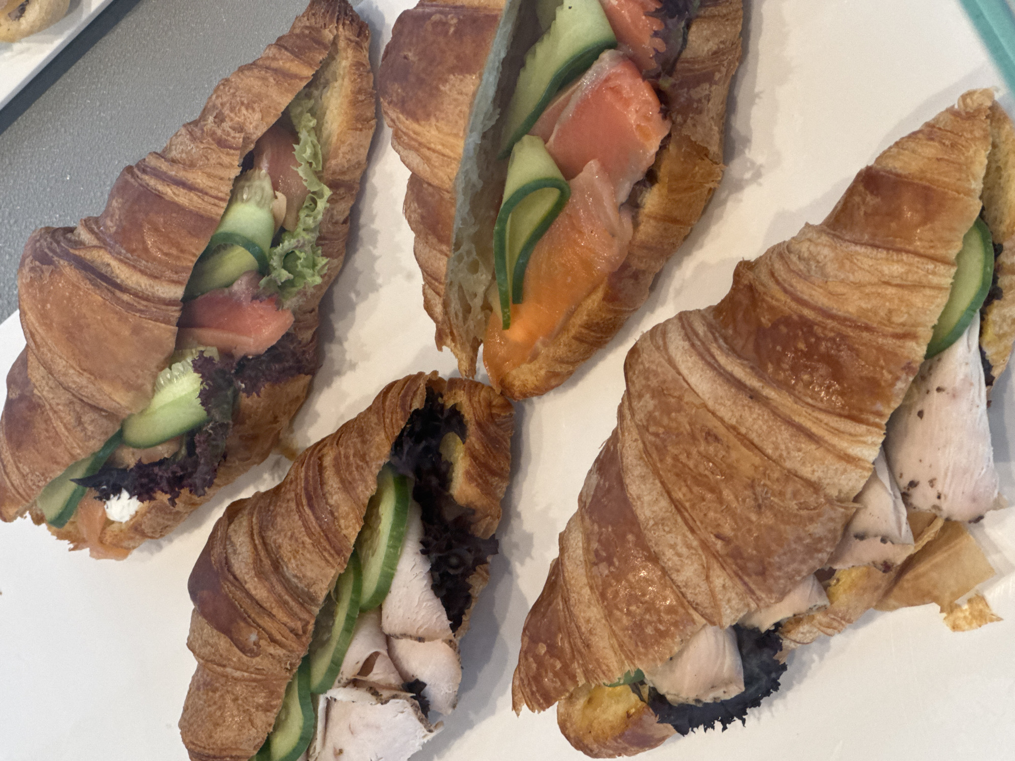 Four croissants, each filled with various ingredients, are arranged on a white surface, possibly a tray or table. Two croissants are filled with sliced turkey, lettuce, and cucumber. One of these turkey croissants is in the foreground, with the turkey visible at the left side and the sliced cucumber on top of the lettuce. The second turkey croissant is positioned behind and to the left of the first, with its contents also visible. The other two croissants contain smoked salmon, lettuce, and cucumber. One salmon croissant is in the background, and another is to the right of the turkey croissants, showcasing the pink salmon slices and green cucumber. The croissants appear to be golden brown and flaky, indicating they are freshly baked. The lighting is bright, highlighting the textures and colors of the food. The background is mostly white with a slight gray element on the right.