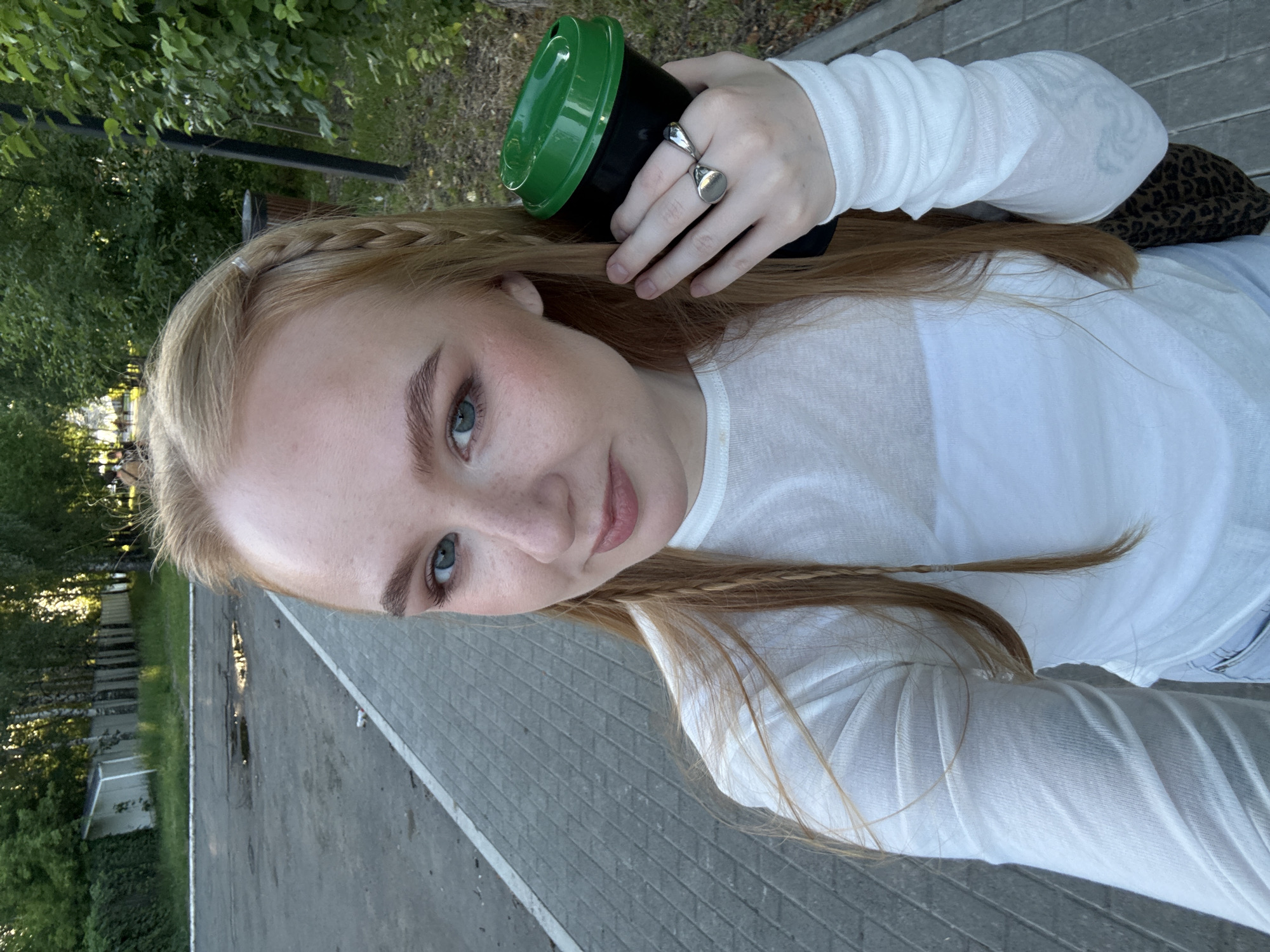 A young woman with blonde hair, styled in braids, is taking a selfie outdoors in Chaikovsky, Russia. She is wearing a white long-sleeved shirt and is holding a black cup with a green lid in her right hand. She has rings on her fingers. The background shows a paved walkway, a grassy area with trees, and a fence. The lighting suggests it is daytime, possibly late afternoon.
