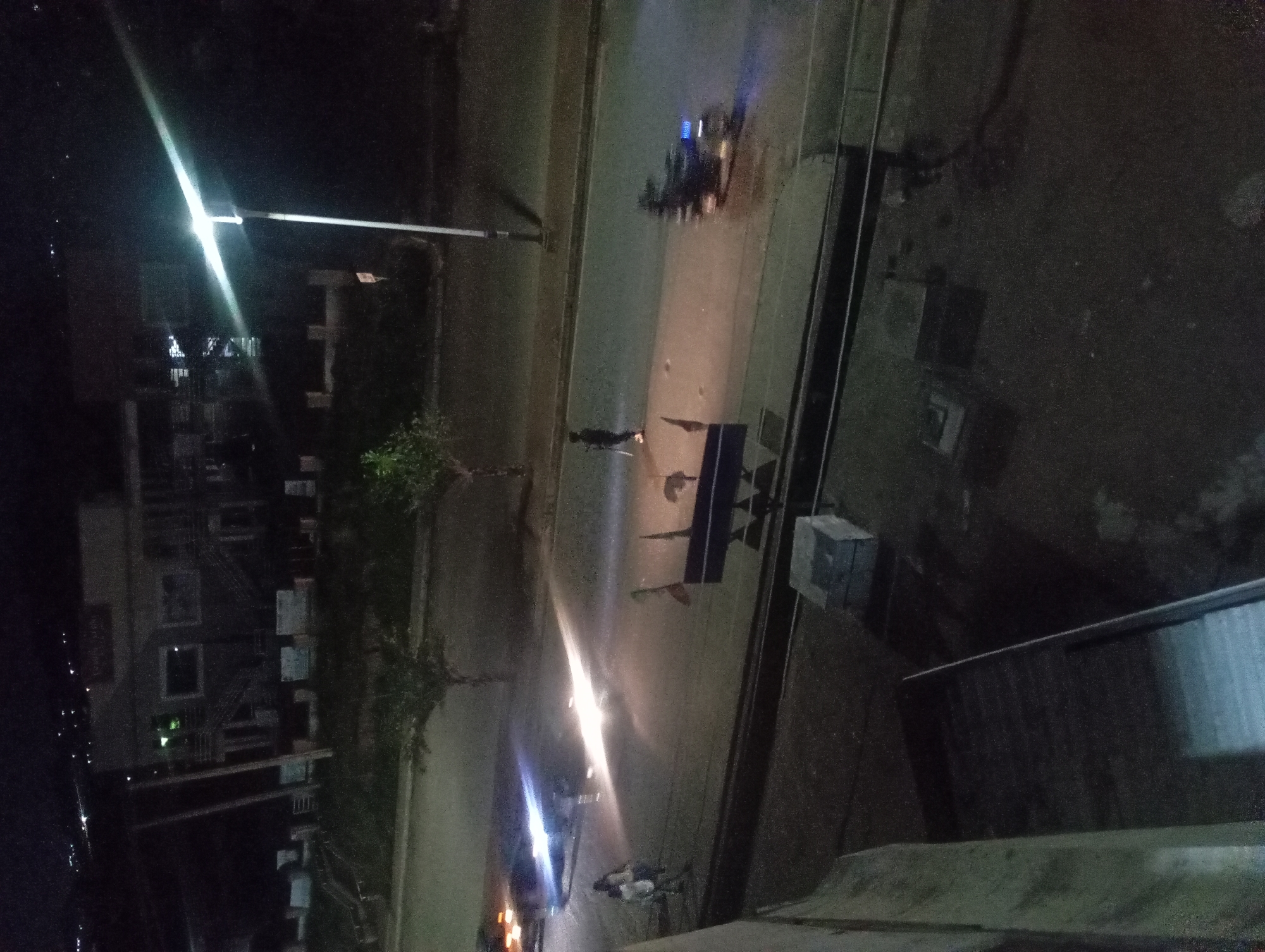A nighttime, high-angle shot captures a street scene in an urban environment. A bright street lamp illuminates a portion of the road. Several vehicles are visible: a car with its headlights on is parked or moving slowly, casting a glare across the road. A motorcycle is also present, with its headlights and possibly taillights illuminated, appearing to be in motion. Two individuals are on the road; one, a pedestrian, is standing in the middle of the road, appearing to hold a long object. Another person is walking on the sidewalk in the background. Buildings, some with lit windows, are visible on the left side of the image. The road surface is asphalt, and there is a sidewalk or paved area adjacent to it. The foreground shows a dark, textured surface, possibly a balcony or rooftop, with some stacked objects. The overall lighting is dim, with the street lamp and vehicle lights being the primary sources of illumination.