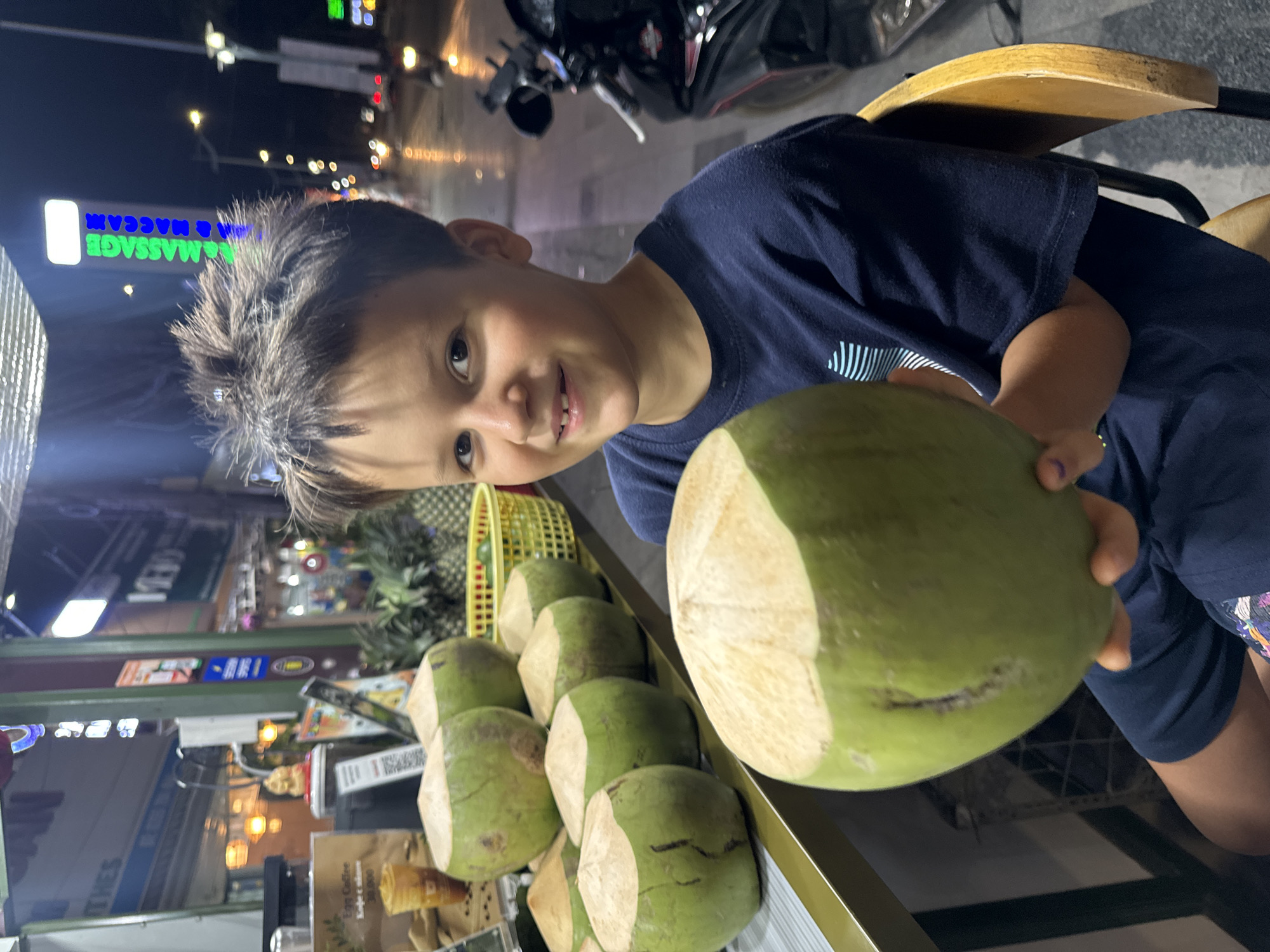 An outdoor, nighttime photograph, rotated 90 degrees counter-clockwise, depicts a young boy holding a fresh green coconut at a street vendor's stall in Mũi Né, Vietnam. The boy, of elementary school age with dark hair and wearing a dark blue short-sleeved t-shirt, smiles directly at the camera, appearing happy and engaged.
He holds a large, light green coconut that has been partially chopped open at the top, ready for drinking. Below and behind his coconut, numerous other similarly prepared green coconuts are neatly displayed on a golden-colored shelf, suggesting a stall specializing in fresh coconut drinks. To the left, a yellow plastic basket holds additional tropical fruits, possibly pineapples.
The background reveals a lively street scene under artificial light. A prominent neon sign, glowing green and blue, displays the reversed text "MASSAGE & MACCAM". Other illuminated signs and streetlights are visible, indicating a commercial area. A motorbike is partially visible parked on the street in the distance. A wooden chair back is seen behind the boy's right shoulder. One discernible sign in the foreground advertises "EGG COFFEE" with a price that appears to be "30.000". The overall ambiance suggests a casual evening outing in a tropical urban environment.