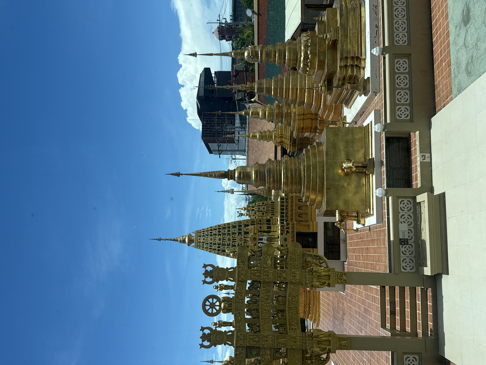 The image shows golden structures, likely part of a temple, under a bright blue sky with some clouds. The structures are ornate, with tiered spires and decorative elements including figures and wheels. A modern building can be seen in the background, suggesting a juxtaposition of ancient and contemporary elements. The location is Bang Pu Subdistrict Municipality, Thailand, based on the architectural style which is typical of Thai temples. No people are visible. The focus is on the detailed golden structures and their relationship to the sky and background buildings.