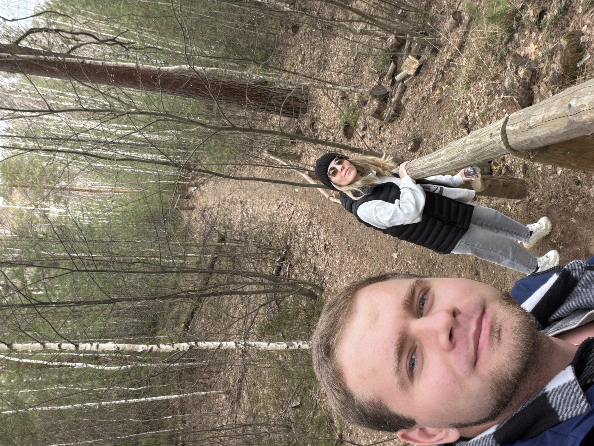 The image is a selfie taken by a man with short blond hair, blue eyes, and a beard, wearing a blue and black plaid jacket. Behind him and slightly to the left, a woman with long blond hair and sunglasses, wearing a black beanie, grey pants, a white sweatshirt, and a black vest is holding onto a wooden handrail. They appear to be in a forest, with bare trees and a leaf-covered ground. The location is Chumna, Russia.