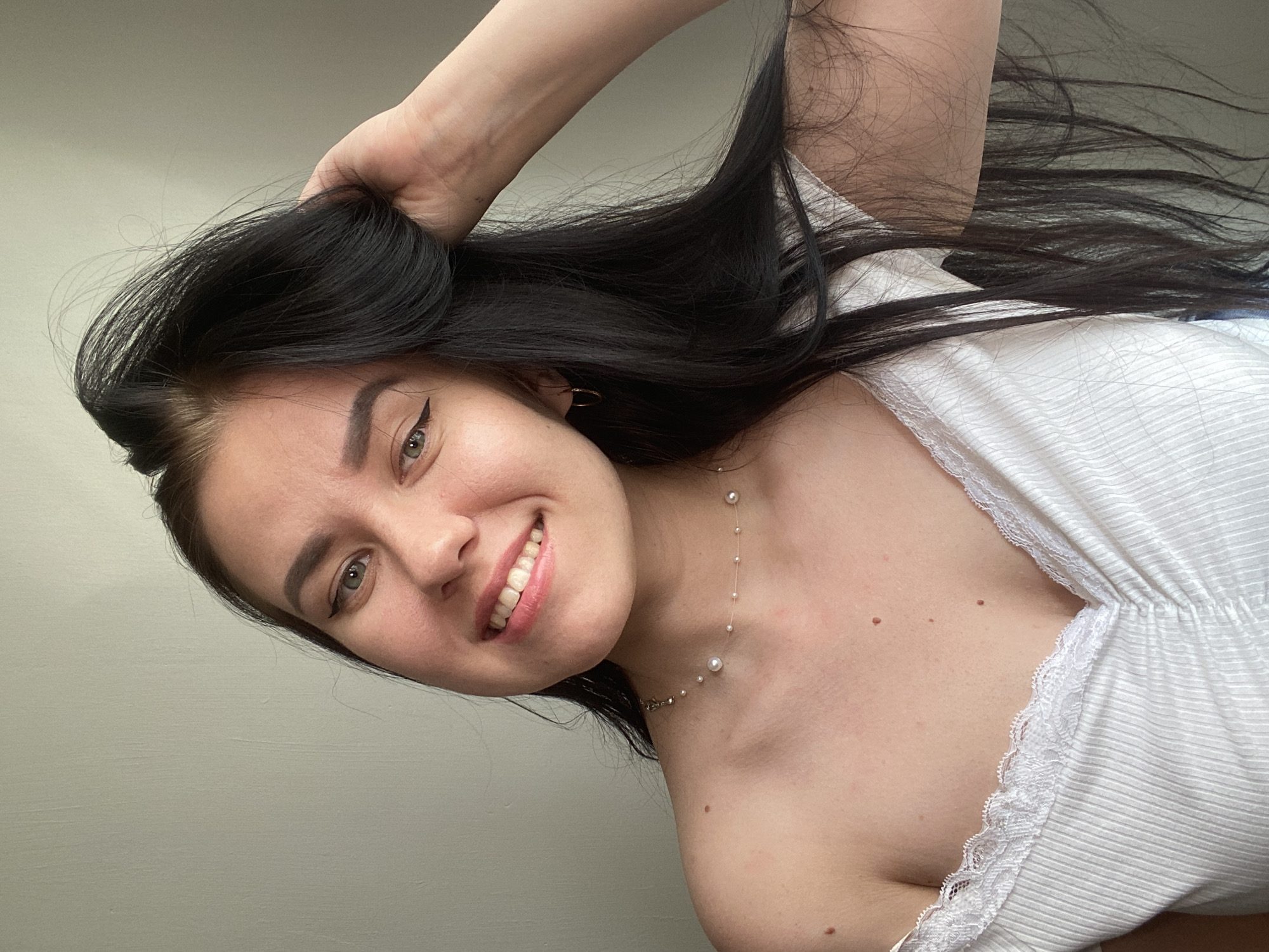 A young woman with long, dark hair is posing for a portrait. She has a light complexion, green eyes, and is wearing a white off-the-shoulder top with lace trim. A delicate pearl necklace adorns her neck. Her right hand is raised, and she is holding her hair. The background is a plain, light-colored wall, suggesting an indoor setting, likely a home environment in Chaikovsky, Russia.