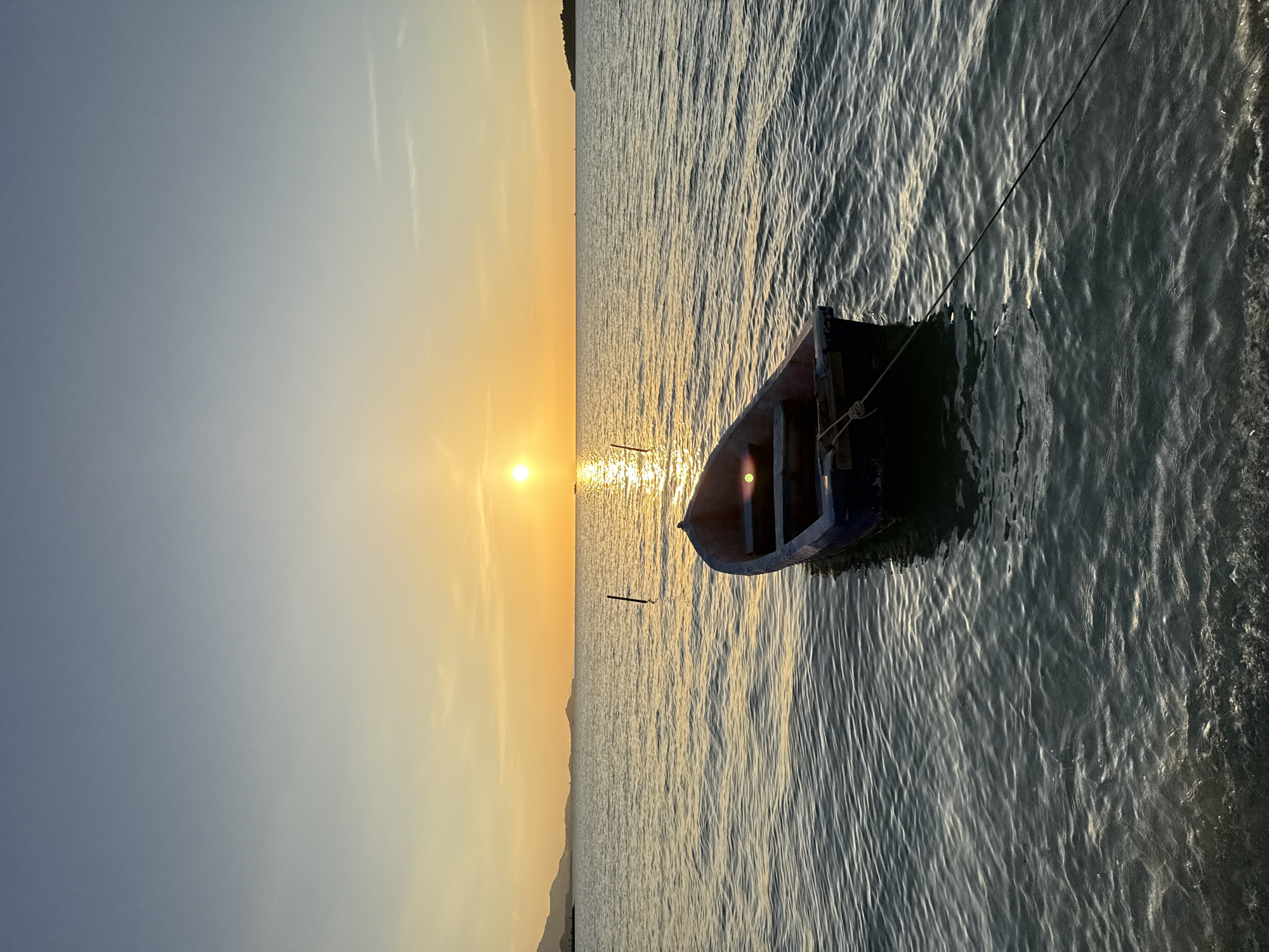 A small blue and red rowboat is anchored in calm water during sunset. The sun is low on the horizon, casting a warm golden glow across the sky and reflecting on the water. In the distance, a coastline with rolling hills is visible. The scene appears to be on the island of Ko Samui in Thailand. The water is clear and tranquil, with gentle ripples on its surface. The boat is tethered to a mooring line, suggesting it is a quiet and peaceful evening. There are no people in the image.