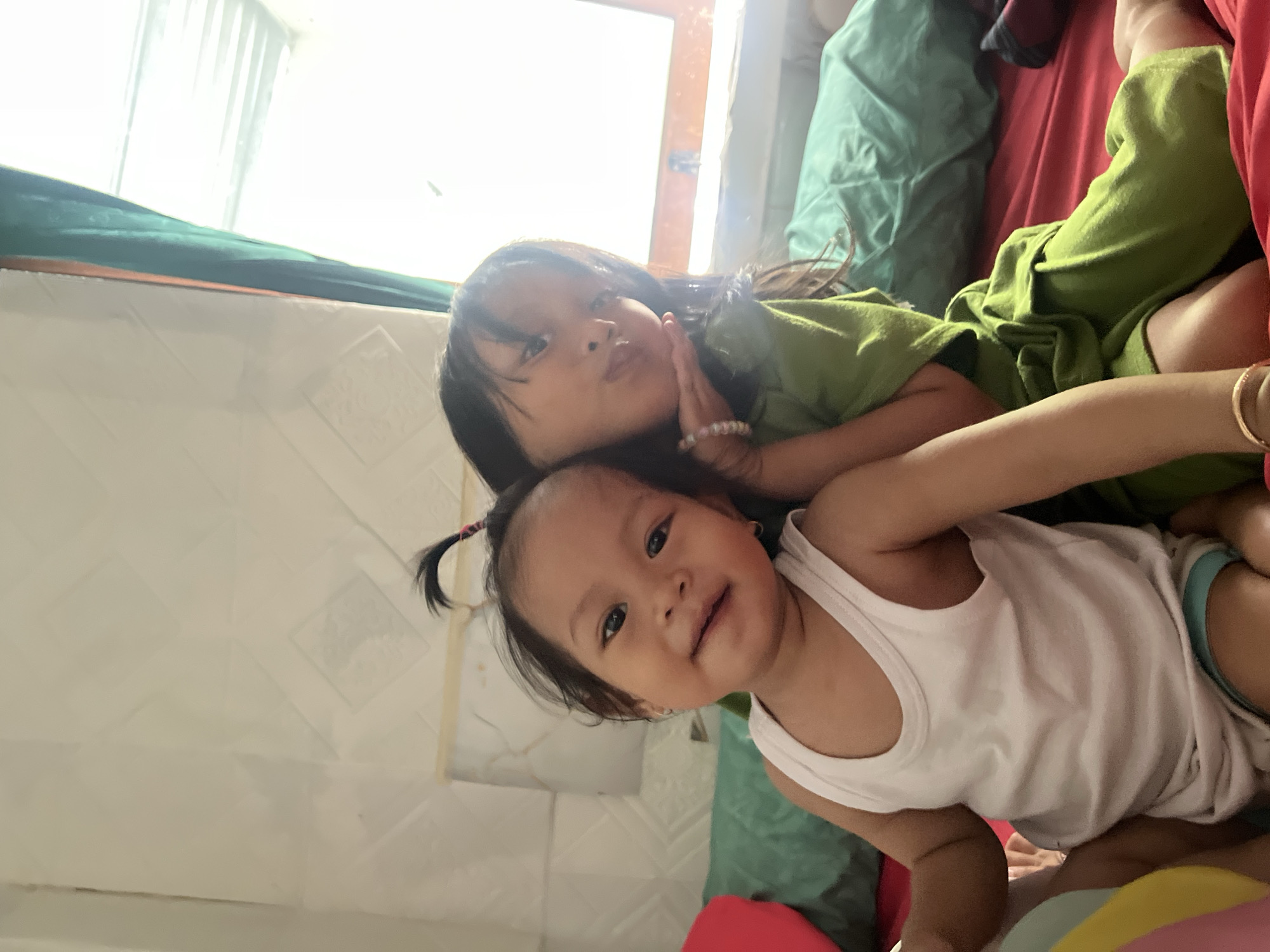 Two young girls are nestled together on a bed. The older girl, on top, is wearing a green t-shirt and rests her head on the younger girl's shoulder. She has dark hair and is looking towards the camera with a slight smile, her hand gently touching her cheek. A beaded bracelet adorns her wrist. The younger girl, beneath, is wearing a white tank top and blue shorts. She looks directly at the camera with a sweet, innocent expression and a faint smile. Her dark hair is pulled into a small ponytail with pink accents. The girls are surrounded by colorful blankets and pillows in shades of green, red, and pink, suggesting a cozy and playful environment. The scene is illuminated by natural light streaming in from a window, possibly indicating daytime. The background suggests an indoor setting, perhaps a bedroom. The overall mood is one of affection and comfort. There is no visible text in the image.