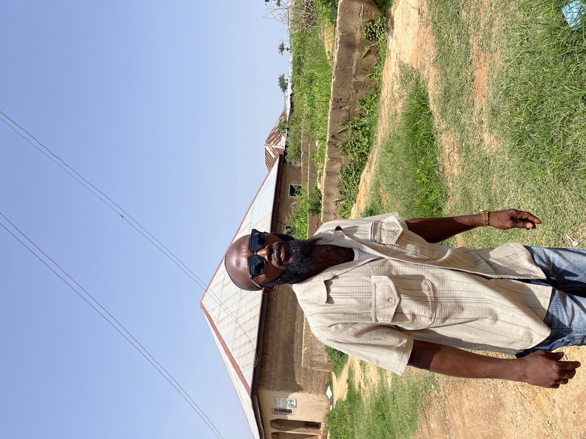In Jos, Nigeria, a man with a beard and sunglasses stands outdoors on a dirt path beside a building with a corrugated metal roof. He is wearing a short-sleeved, striped button-up shirt and blue jeans. The background shows more buildings with similar roofs and lush green vegetation. Overhead, power lines stretch across a clear blue sky. The man is looking towards the camera, and his left arm is bent at the elbow, while his right arm is extended downwards. A gold watch is visible on his left wrist. The building behind him has a tan-colored exterior and a window.