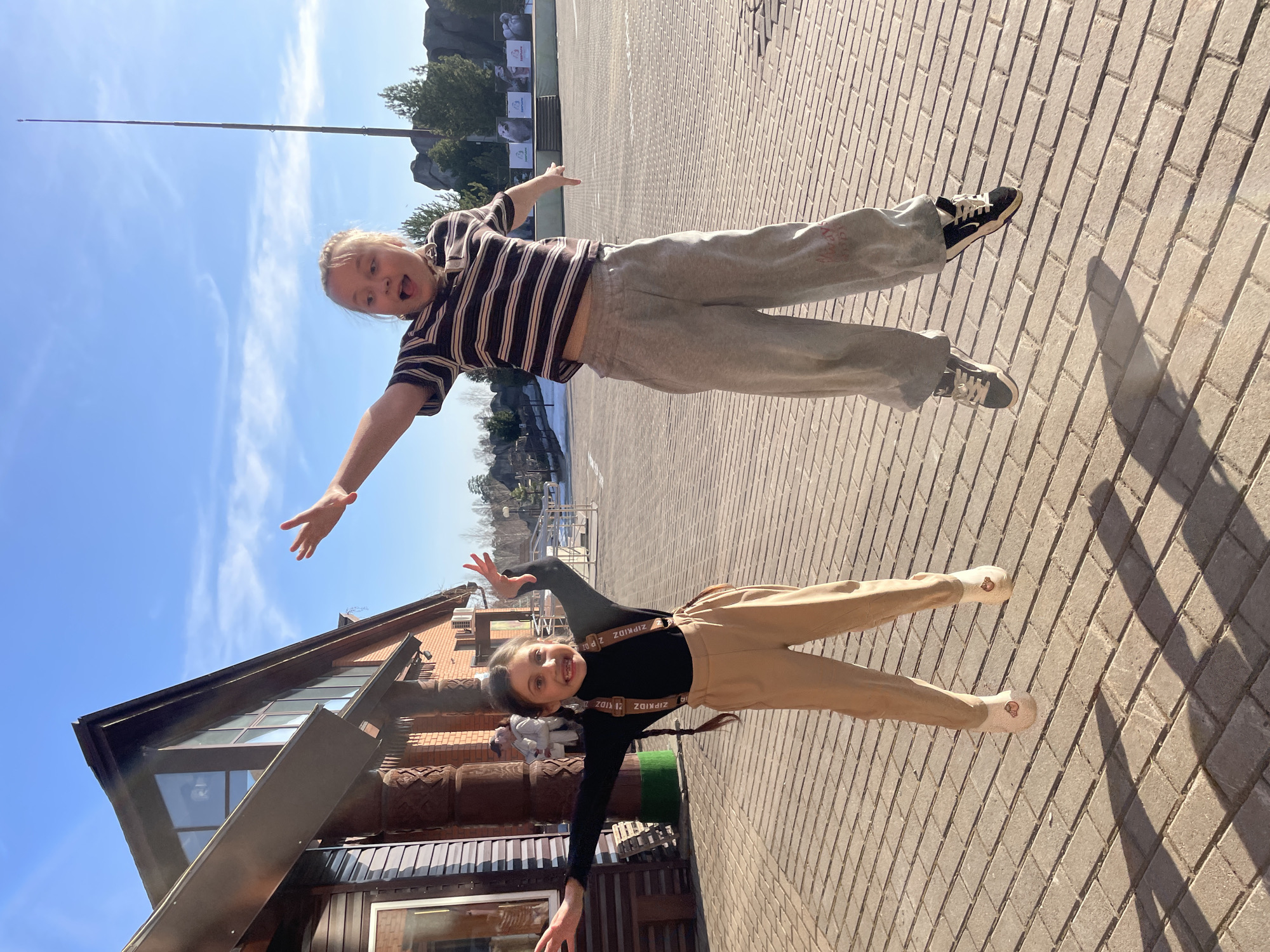 Two girls are photographed outdoors on a paved surface. The first girl, positioned above in the image, is wearing a brown and white striped shirt and gray sweatpants with sneakers. She has her arms outstretched and her mouth open. The second girl, below, is wearing a black long-sleeved shirt, tan pants, and light-colored shoes. She also has her arms outstretched. In the background, there is a building with windows and architectural details, as well as trees and a blue sky. The scene is likely in Izhevsk, Russia.