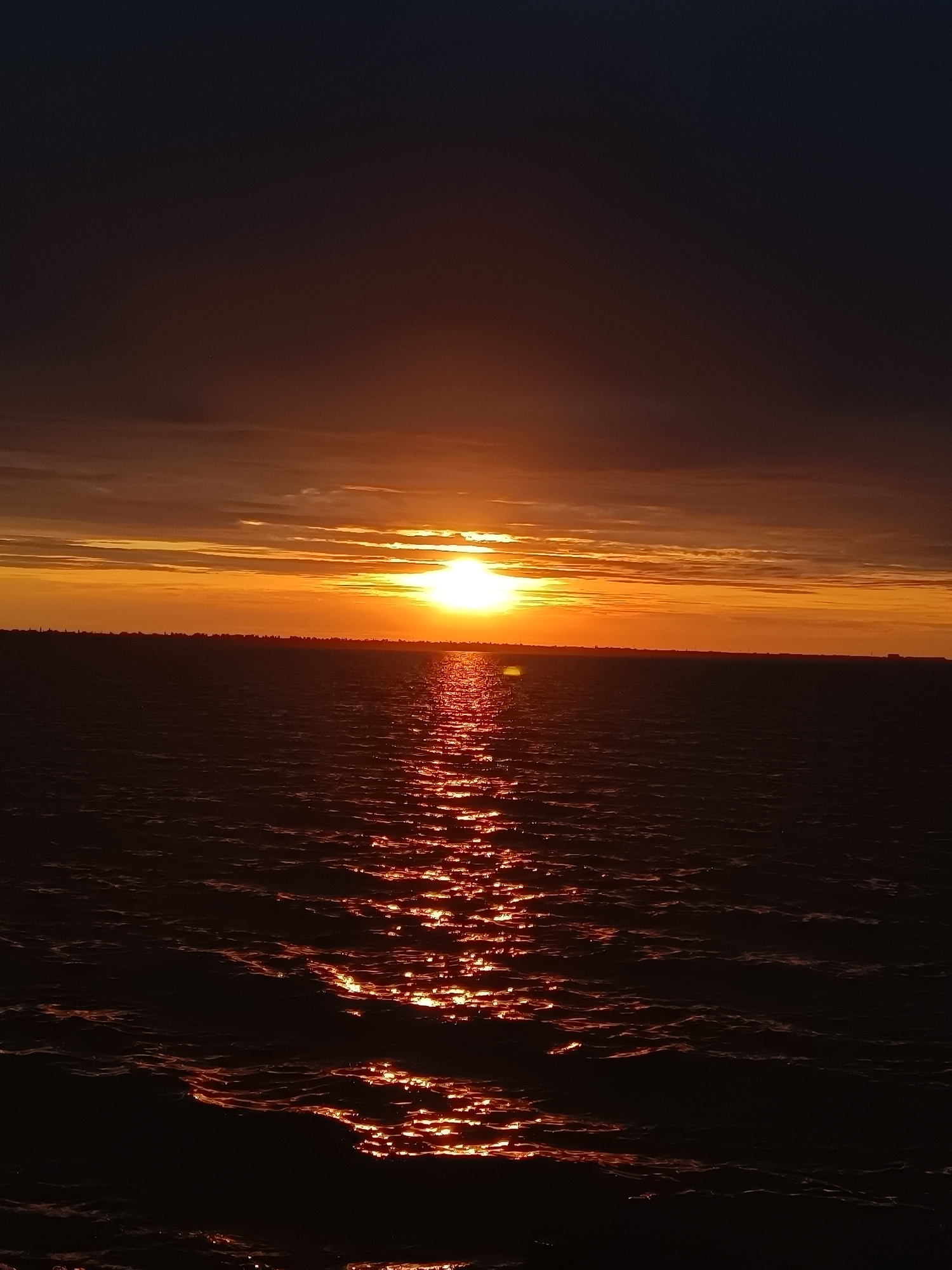 A vertical shot captures a sunset over the Black Sea near Zatoka, Ukraine. The sun, a bright orange orb, is positioned in the upper center of the frame, casting a shimmering reflection across the dark, choppy water. The sky transitions from a deep indigo at the top to vibrant orange and yellow hues near the horizon, with wispy clouds streaking across. A faint, dark silhouette of land is visible along the horizon line, suggesting the presence of buildings or trees. The water's surface is characterized by small waves, each catching the sunset's light and creating a dazzling, textured pattern. The overall scene is one of natural beauty and serene twilight ambiance.