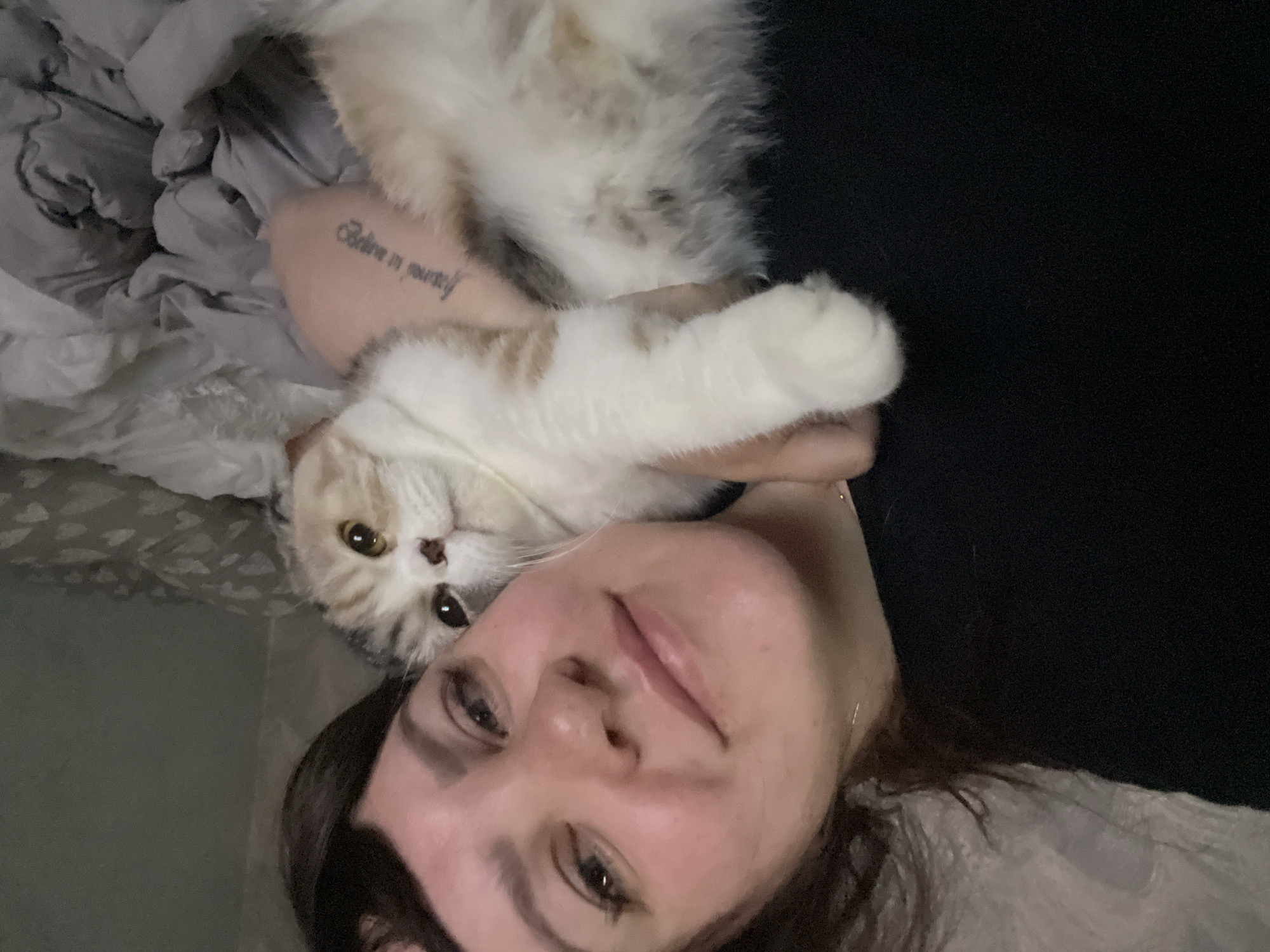 A close-up, intimate portrait features a person and a fluffy cat sharing a moment of calm within an indoor setting. The image is taken from an overhead perspective, with the person's face occupying the lower half of the frame. They have dark hair and a soft, relaxed expression, looking directly at the camera with a slight, gentle smile.
Resting comfortably on the person's arm or chest is a domestic cat, primarily white with distinct light brown or tan patches, particularly around its head and back. The cat also looks directly towards the camera, appearing calm and content. A notable detail is a tattoo on the person's arm, which reads "Believe in yourself" in a flowing script font.
The background consists of soft, crumpled fabric, predominantly light grey and white, suggesting bedding or a comfortable blanket, indicating the scene takes place on a bed or sofa. The right side of the image features a dark, undefined area, possibly a dark blanket or a dimly lit wall. The lighting is soft and ambient, characteristic of an indoor environment, potentially in the evening or during low light conditions. The overall mood of the image is one of affection, peace, and companionship between the individual and their pet. The location context for this image is Noviy, Russia.