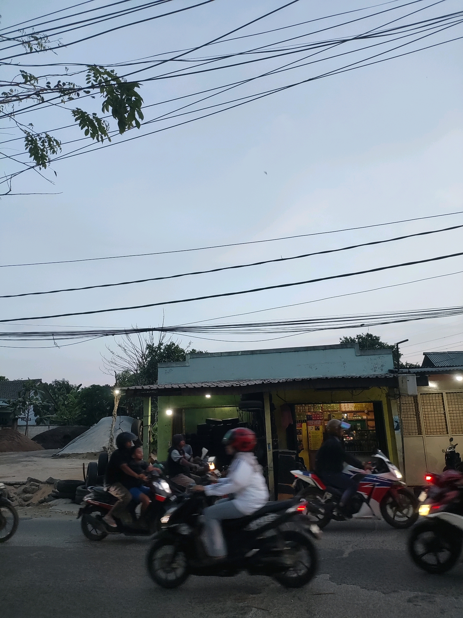 The image depicts a street scene in Wanasari, Indonesia, at dusk or early morning. The sky is a pale blue, overcast with numerous power lines strung across the frame. Vegetation, including leafy branches, encroaches on the power lines from the upper left. In the mid-ground, a low-rise building with a light blue facade and an open-fronted area displays shelves stocked with goods, suggesting it is a shop or warung. Two streetlights are visible, one near the building and another further back among trees.
The foreground is dominated by a paved road where several motorcycles are in motion. The blur of the motorcycles indicates movement. On the leftmost motorcycle, the rear wheel is partially visible. To its right, a motorcycle carries two individuals: a person in the front wearing a black helmet and a child behind them wearing a blue shirt. Further right, another motorcycle is ridden by a person in a white jacket and a red helmet, their face obscured. Another motorcycle, predominantly red and white, is ridden by a person wearing a dark jacket and a blue cap. The rear of this motorcycle shows a red taillight. In the far right, the front of a red motorcycle is visible, its headlight illuminated.
The background includes more vegetation, houses with tiled roofs, and a pile of sand and tires on the left side of the road. The overall impression is of a busy, active street in a residential or commercial area of Wanasari.