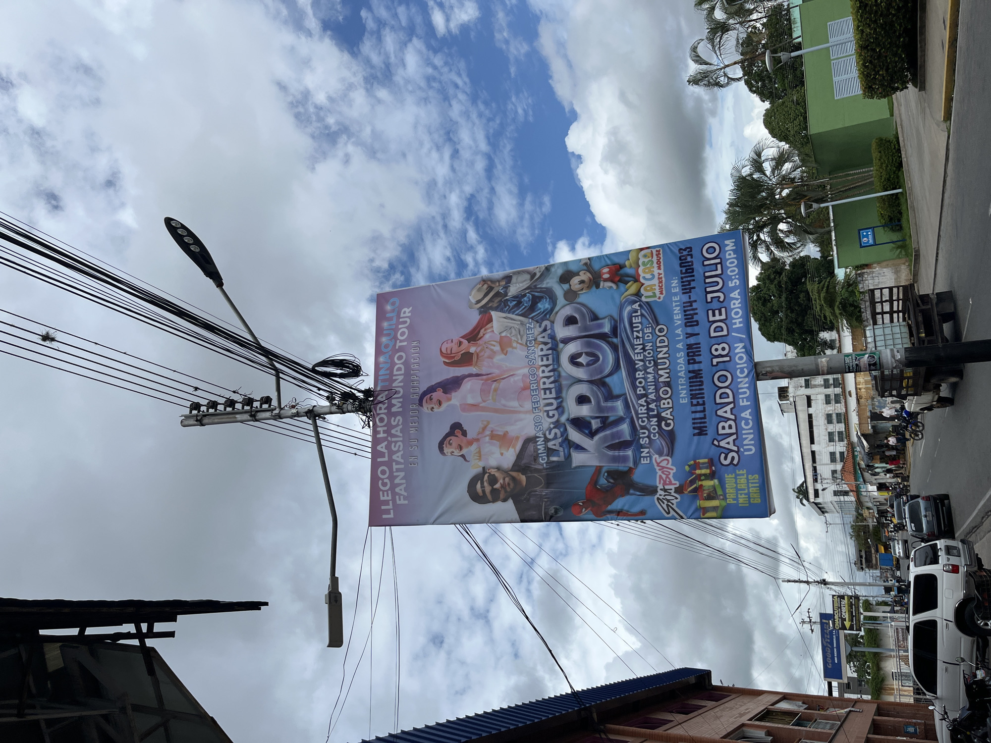 A horizontal medium shot depicts a street scene in Tinaquillo, Venezuela, dominated by a large advertising banner stretched between utility poles. The banner promotes "KPOP" performances at the Gimnasio Federico Sanchez-Las Guerreras, with shows on Saturday, July 18, at 5:00 PM. The banner features cartoon characters, including Mickey Mouse and Spiderman, alongside real people. The banner advertises a "TINAQUILLO FANTASIAS MUNDO TOUR".
Utility poles with numerous black wires run across the top of the image, partially obscuring a cloudy blue sky. Below, street-level activity includes parked cars and motorcycles, and a pedestrian. A green building and other structures are visible on the right side. The image is slightly off-kilter, giving the impression of a tilted perspective.