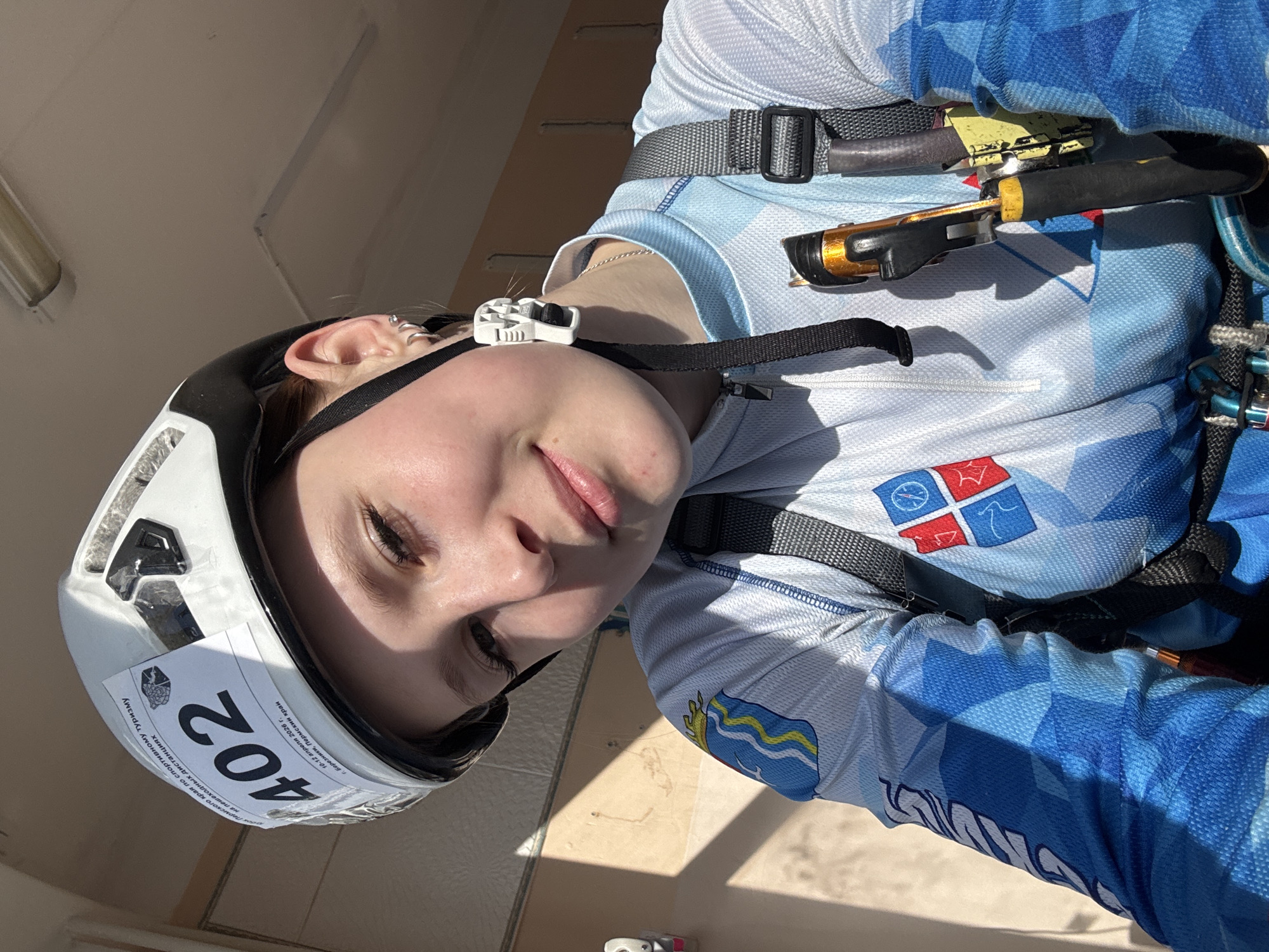 A young woman is wearing a white climbing helmet labeled with the number "402" and text in Russian, which translates to "on pedestrian distances of the Perm region on sports tourism. Berezniki, Perm region 10-12 April 2026." She is also wearing a blue and white patterned athletic shirt and climbing gear. The location is Berezniki, Perm region, Russia. The event took place on April 10-12, 2026.