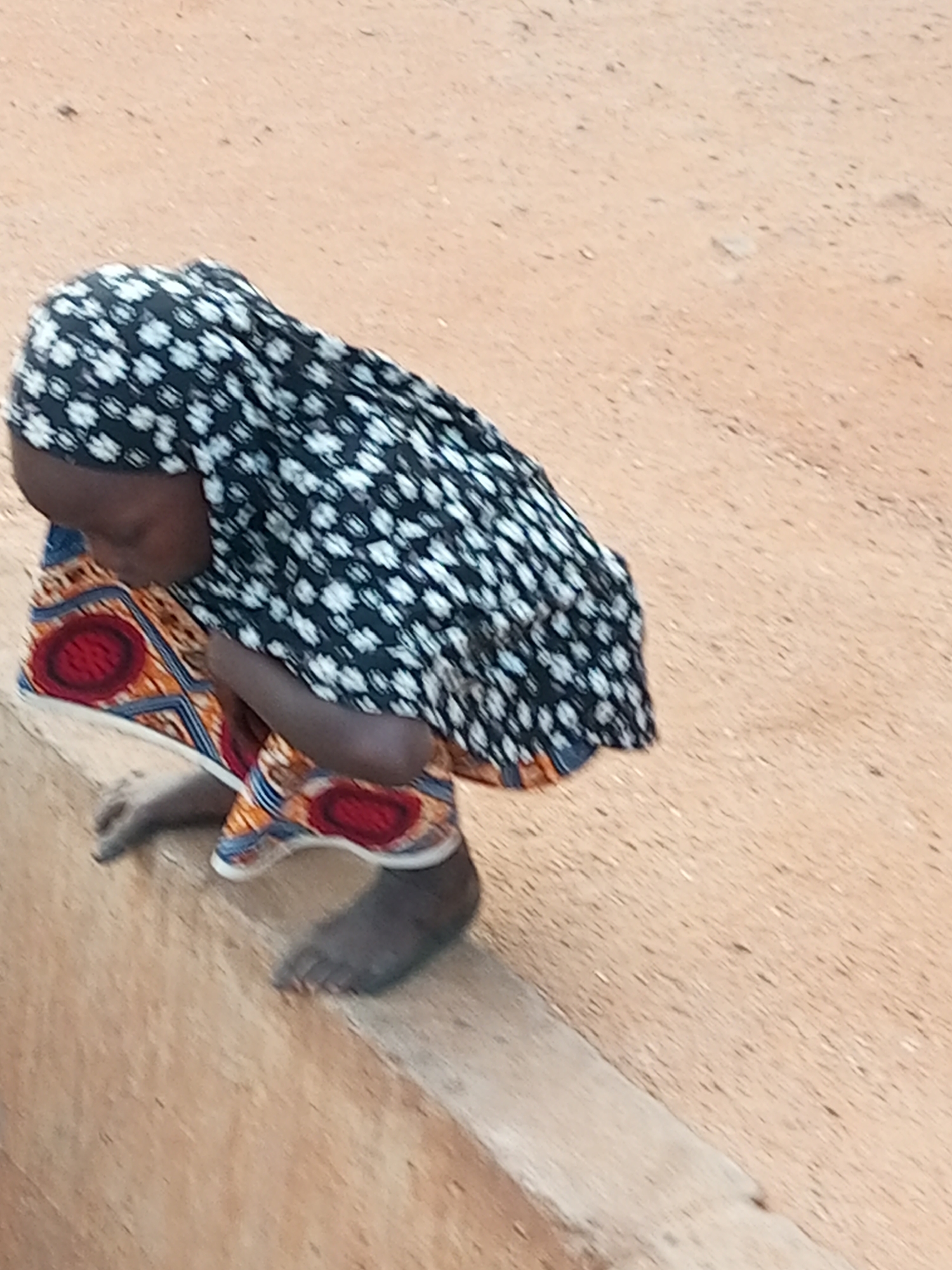 A young person, likely a child, is crouching on a dirt ledge. They are wearing a black and white patterned headscarf and a colorful garment with red circular patterns. Their bare feet are visible, resting on the ledge. The ground around them is sandy or dusty. The scene appears to be outdoors, possibly in a rural or less developed urban area. The image does not contain enough information to definitively place it in Gombe, Nigeria, or any other specific city.