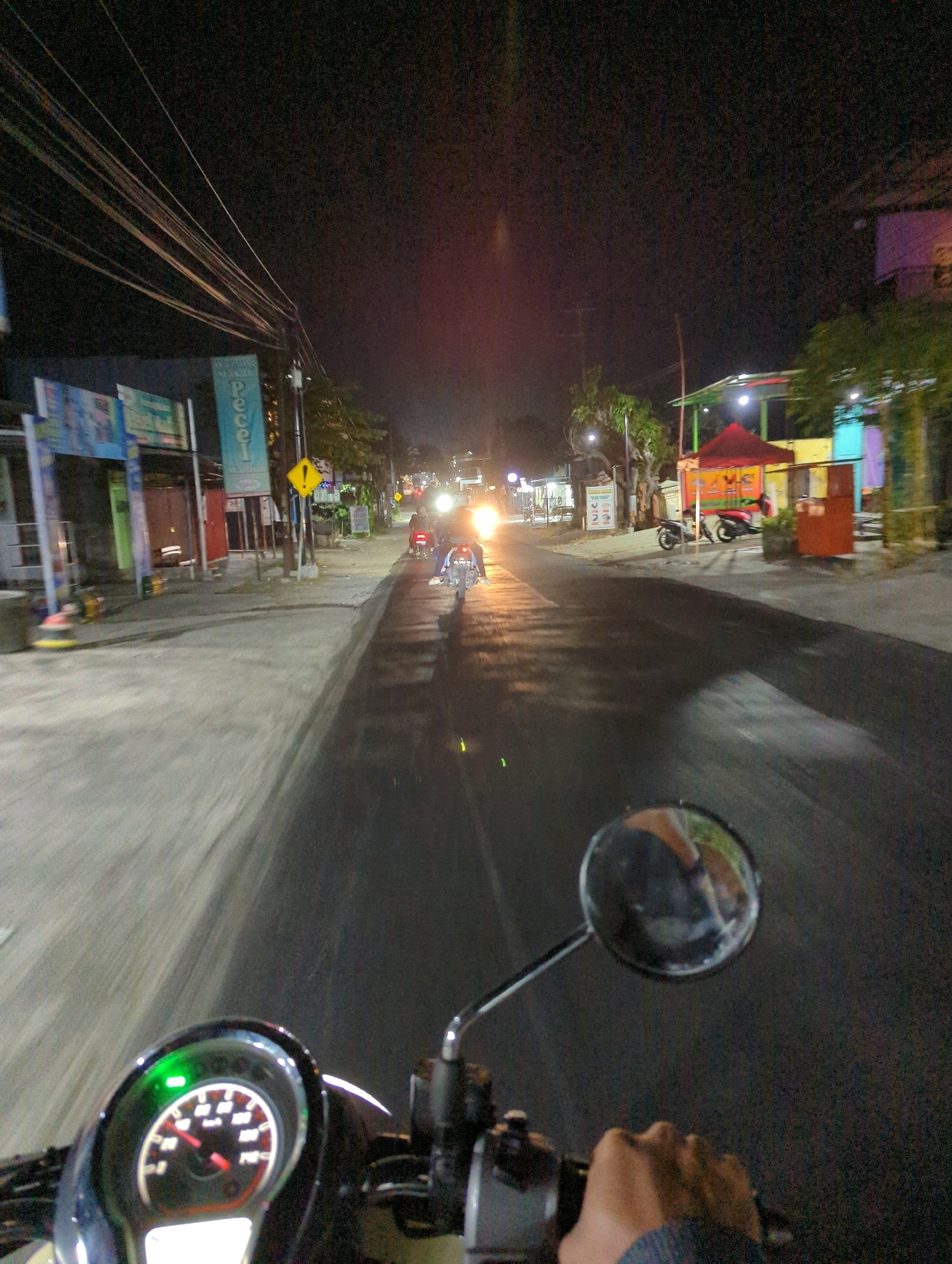 The image captures a first-person perspective of someone riding a scooter at night on a street in Kacangan, Indonesia. The road is wet and reflects the headlights of passing vehicles and streetlights. On the left side of the road, there are buildings with signs, one of which appears to advertise "Pecel," a traditional Indonesian salad. Power lines crisscross overhead, illuminated by the ambient light. Further down the street, other scooters and motorbikes can be seen, some with their headlights on. The rider's hand is visible on the handlebars, and the scooter's speedometer shows a speed between 20 and 40 km/h, with a green indicator light illuminated. A round rearview mirror reflects a blurred image of the rider and their surroundings. The overall atmosphere is that of a lively street scene at night.