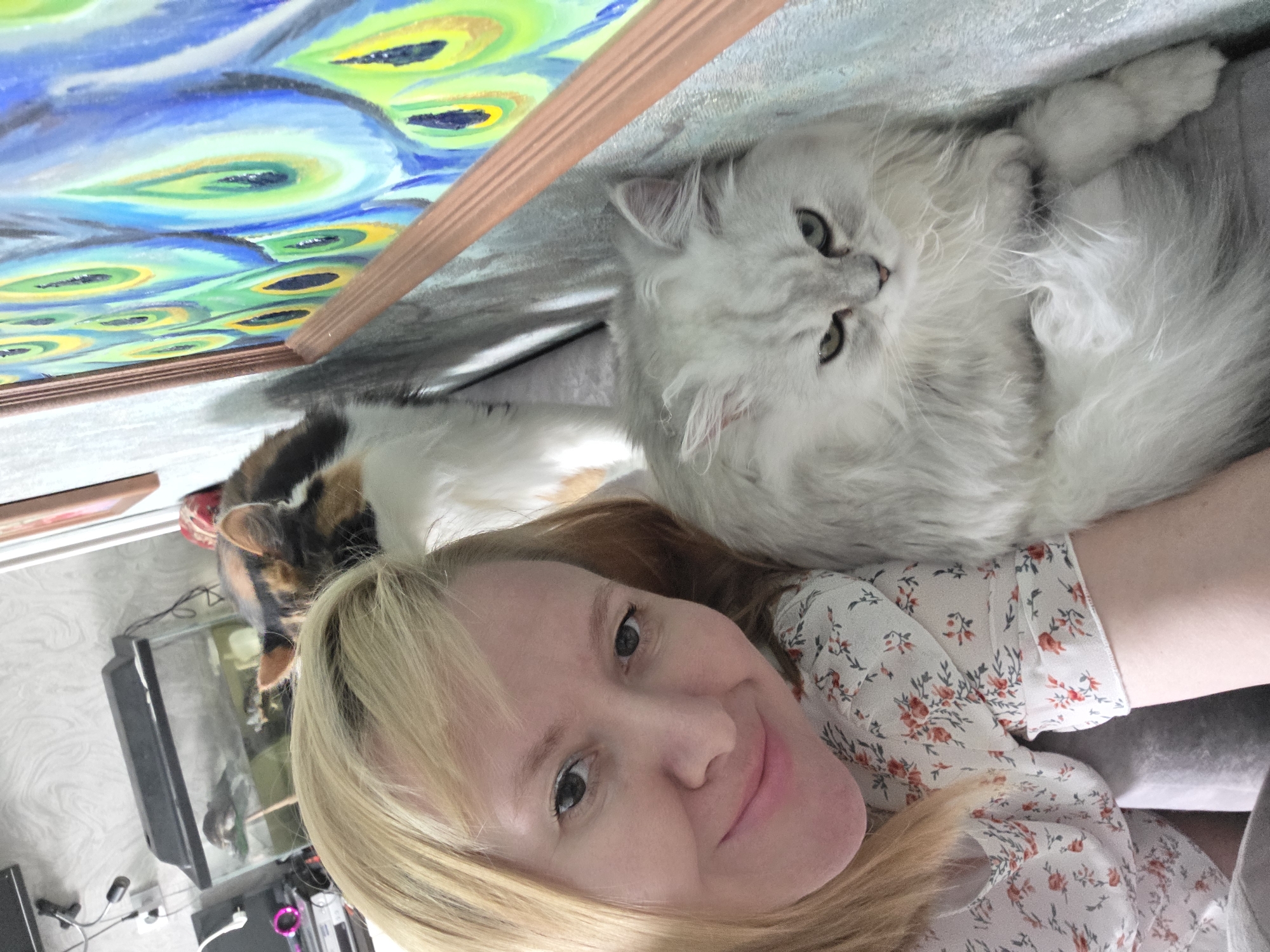 This media file is a close-up selfie taken by a person with blonde hair, smiling at the camera. They are wearing a white blouse with a floral pattern of red and green. To the right of the person, a fluffy white cat is lying down, with its head turned towards the camera. Behind the cat and to the left, another cat with calico fur is partially visible, lying on top of what appears to be an aquarium. In the background, a portion of a painting featuring peacock feathers in vibrant blues, greens, and yellows is visible. The overall setting appears to be indoors, possibly a living room, with some electronic equipment and a wall with a textured pattern in the background. The lighting suggests it is daytime. The image is oriented sideways.