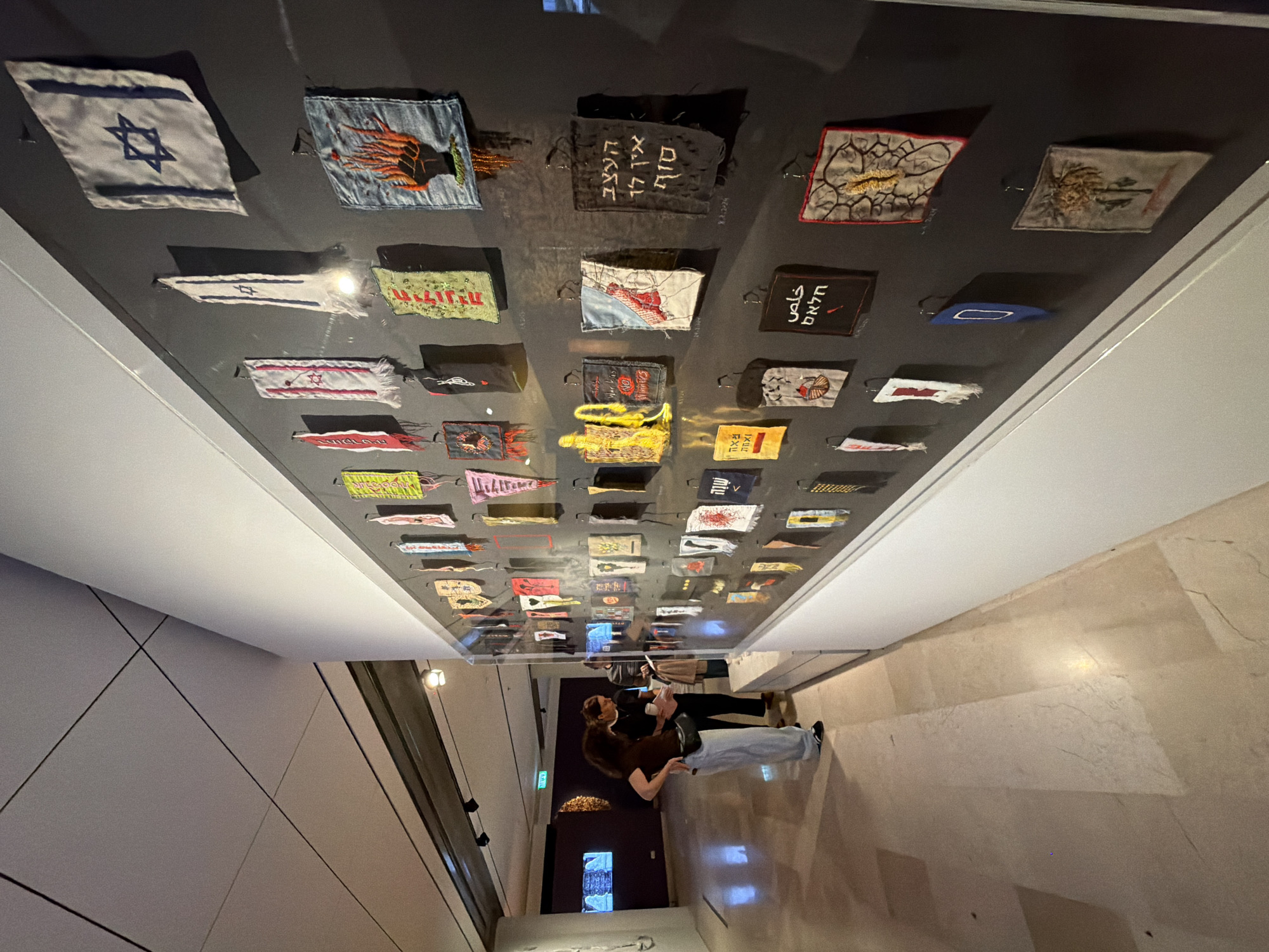 A wide-angle, tilted photograph shows a hallway leading to an exhibit of stitched fabric squares under glass, presumed to be in Tel Aviv, Israel, given the presence of Israeli flags. Two people are in the hallway, one with long brown hair and a dark brown jacket, and the other with shorter, dark hair and a light-colored jacket. The person with long hair is partially obscured by the person with short hair. Both appear to be looking at the exhibit and holding objects that could be pamphlets or phones. The exhibit consists of many small fabric squares stitched with images and text. Several squares feature the Star of David, while others depict various symbols and writings. The lighting is dim, with reflections on the glass covering the fabric squares. The hallway walls are smooth and neutral-colored, while the floor is a light marble or stone. There is a partially visible dark-colored door at the far end of the hall, with a display visible through the opening.