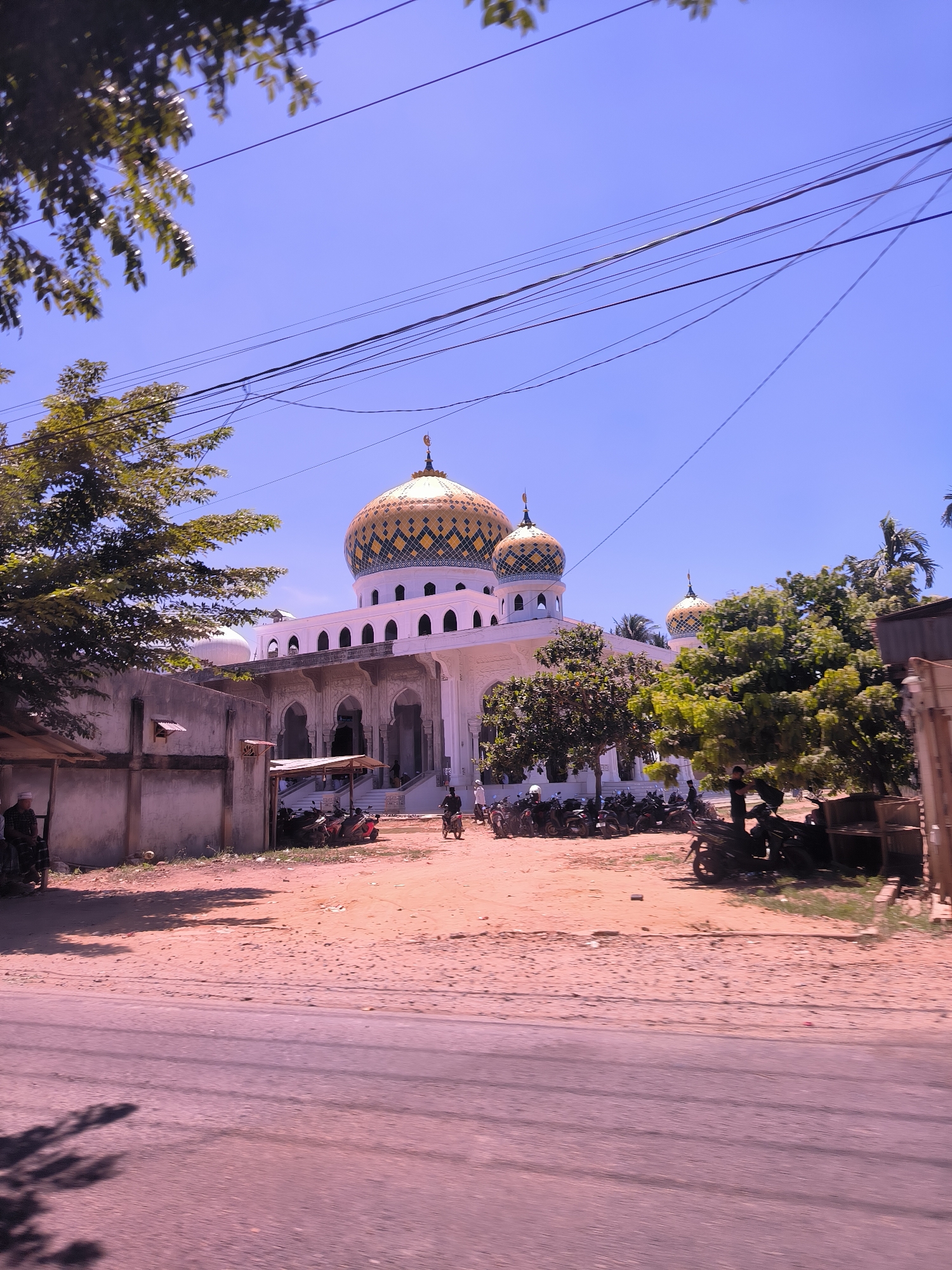 The image depicts a mosque in Keudealue Ie Puteh, Indonesia, under a clear blue sky. The mosque is a white structure with multiple domes, the main one being large and adorned with a gold geometric pattern. There are trees on both sides of the mosque. In front of the mosque, on the ground between the building and the road, are several motorcycles parked. To the left of the mosque is a weathered concrete wall. A person is standing beside this wall. A person is seen driving one of the motorcycles. On the right are some other persons sitting under a tree in front of some wooden furniture. The ground is a mix of dirt and concrete. Power lines stretch across the upper part of the scene. The image is taken from a slightly low angle, suggesting the photographer is on the ground or in a vehicle.