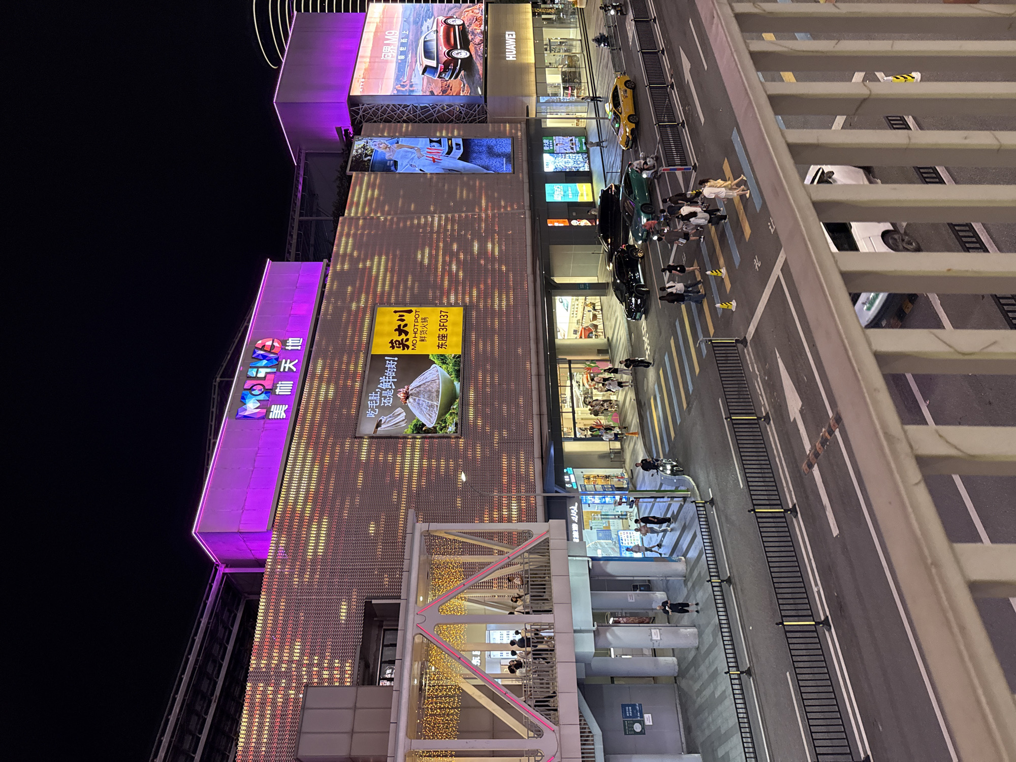 A night view shows the exterior of a large, modern shopping mall in Tianhe District, China. The building is illuminated with bright purple and pink lights along its upper edges and on a prominent sign that reads "MOLT TIME". Large LED screens display advertisements, including one for "MO HOT POT" featuring a close-up of food, and another for an SUV. A pedestrian bridge with decorative string lights connects to the mall, and several people are visible on and around it. Below, a street is busy with traffic, including several cars (a black SUV, a green sedan, a yellow taxi) and a motorcycle. Pedestrians are crossing the street at a marked crosswalk, which is illuminated with yellow and blue stripes. The overall scene depicts a vibrant urban commercial area at night, with prominent signage, illuminated architecture, and street-level activity.