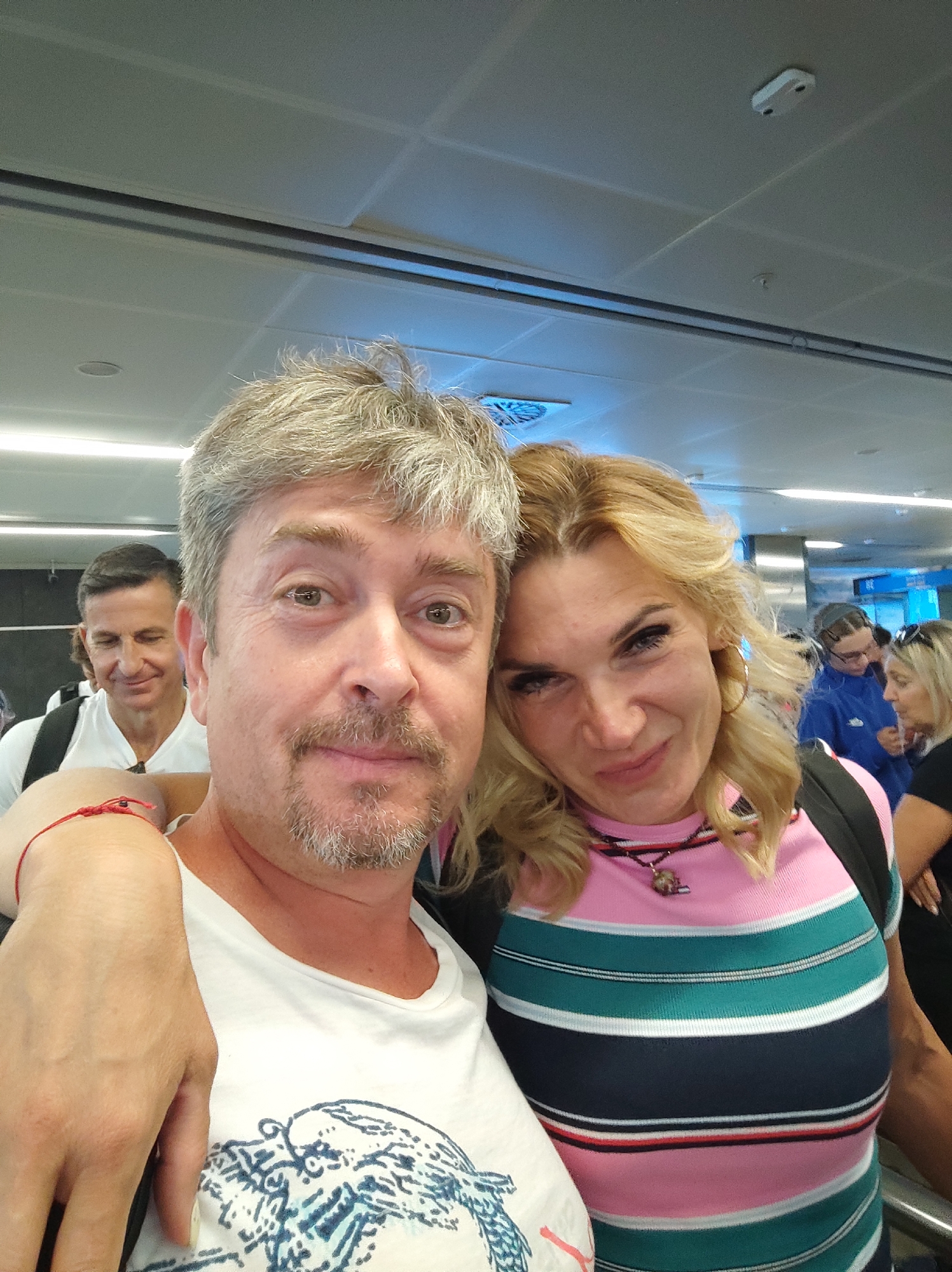 A selfie taken indoors, possibly in an airport terminal, shows a man and a woman posing together. The man in the foreground has graying hair, a mustache, and a goatee. He is wearing a white t-shirt with a graphic design and has his left arm around the woman. The woman next to him has blonde hair and is wearing a striped shirt with pink, teal, white, and navy blue stripes, along with a necklace. Another man is visible in the background, smiling and looking to the left. The environment appears to be a public indoor space with a tiled ceiling and fluorescent lighting. The woman is wearing a red string bracelet on her left wrist. The scene is likely in the city of Beyoğlu, Turkey, given the context of the image.