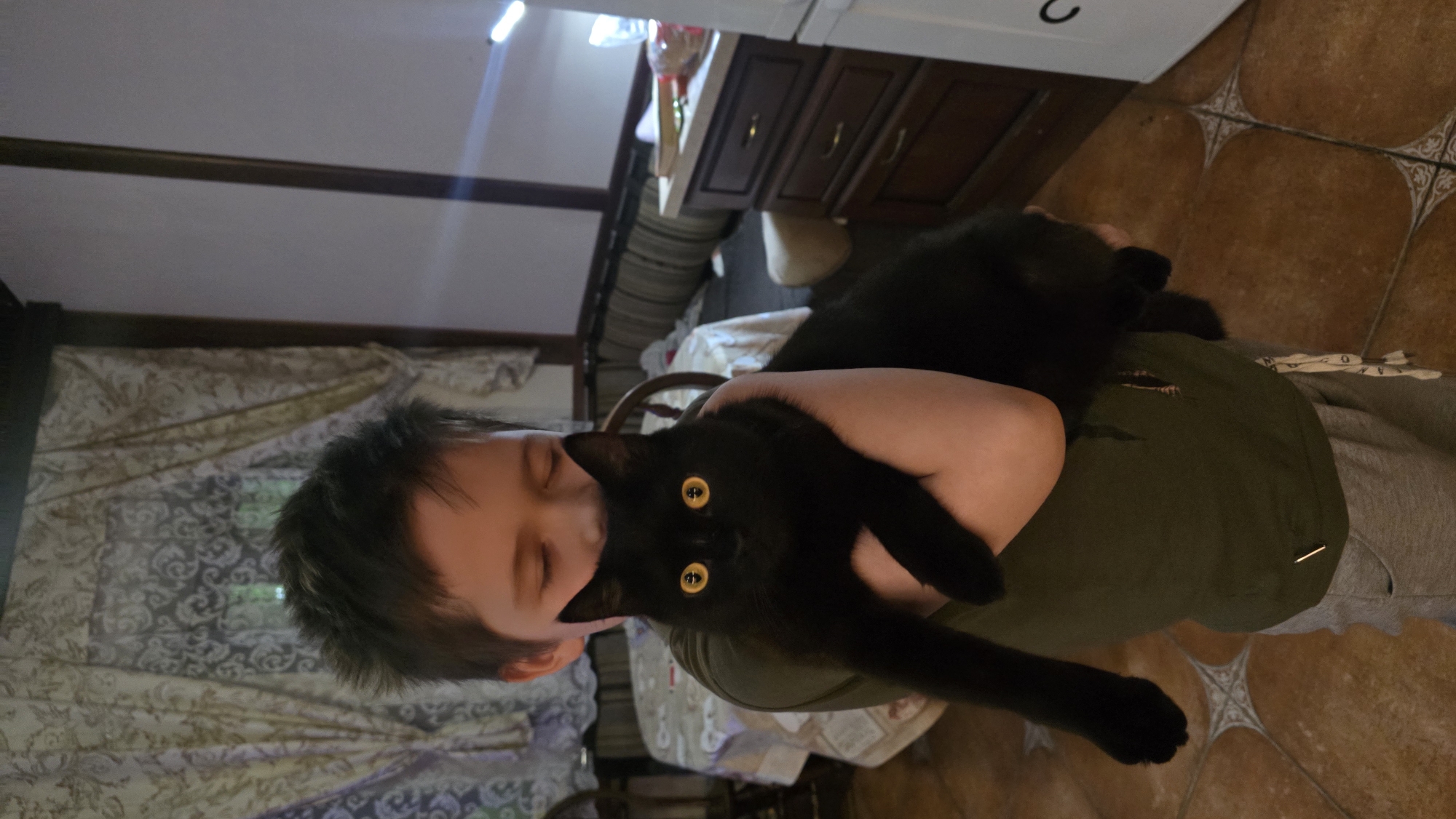 In a dimly lit room, a young boy with dark, spiky hair is holding a black cat with striking yellow eyes. The boy, wearing a dark green t-shirt and grey sweatpants, has his eyes closed and appears to be looking down at the cat cradled in his arms. The cat, with its fur appearing sleek and dark, looks directly at the camera. The background suggests an indoor domestic setting, possibly a kitchen or dining area, with patterned curtains visible behind the boy and a table with a tablecloth to his left. To the right, a wooden cabinet with drawers and a white appliance are partially visible, indicating the scene is likely located within a residence in Il'inskiy, Russia. The floor is tiled with a reddish-brown hue.