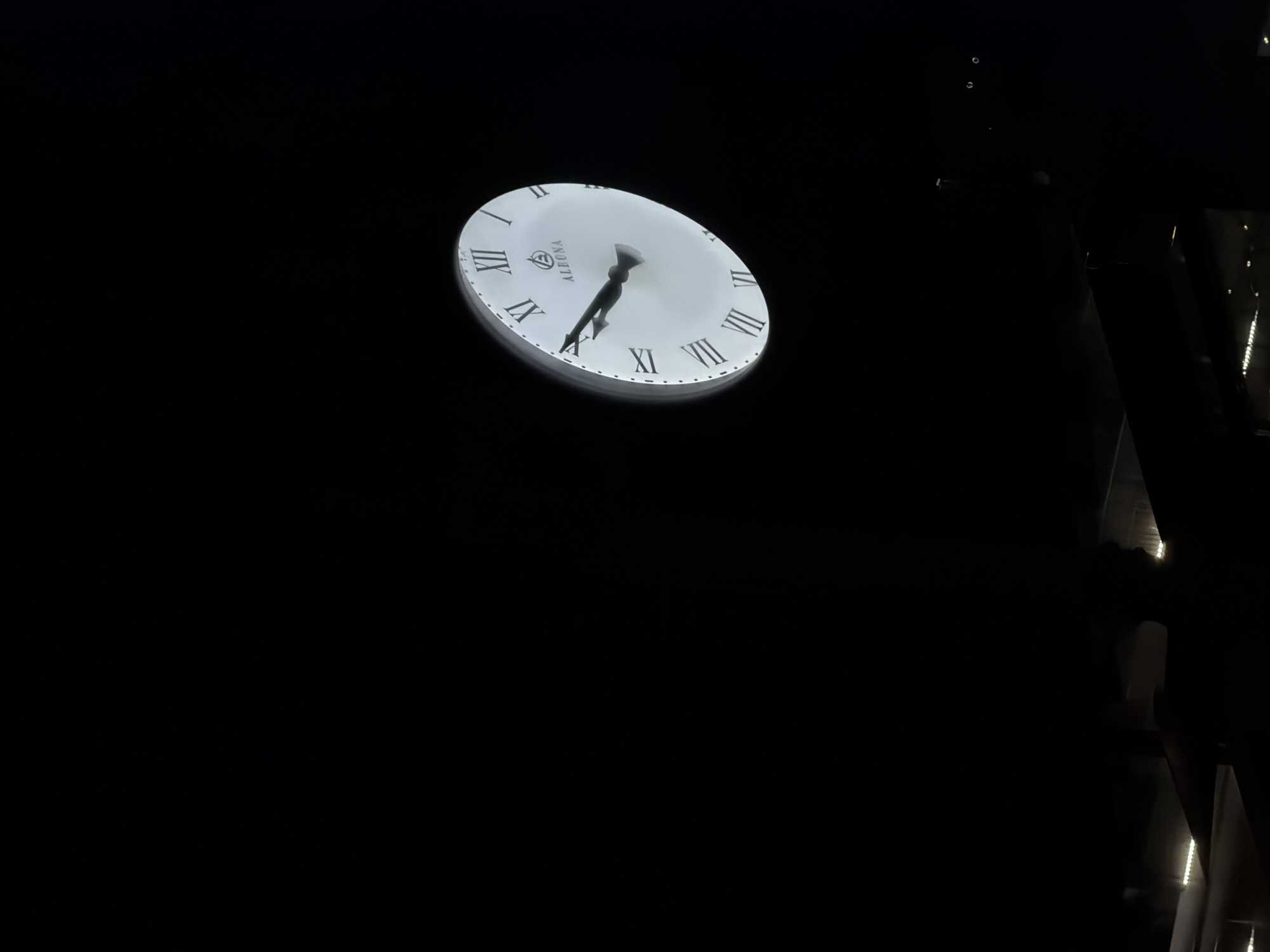 A low-angle shot reveals a white clock face set against a dark background. The clock shows Roman numerals, and its hands indicate approximately 7:30. The name "ALEKNA" is printed on the clock face. To the right of the clock, a dark structure is partially visible, with what appear to be illuminated strips or reflections. The location is Varna, Bulgaria.