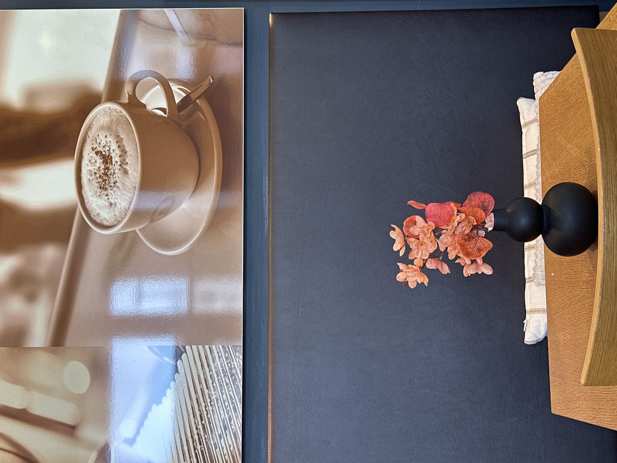 The image shows a close-up of a table setting in what appears to be a cafe or restaurant. In the foreground, a small black vase holds a floral arrangement of peach and red artificial flowers. Behind the vase, a textured pillow with horizontal stripes is visible. The table itself is made of wood with a diagonal grain pattern. In the upper portion of the image, a framed picture or mural depicts a coffee cup with foam and a spoon on a saucer, set against a blurred background. The overall scene suggests a comfortable and decorative dining environment. The provided information indicates the location is in Krefeld, Germany. No people are present in the image.