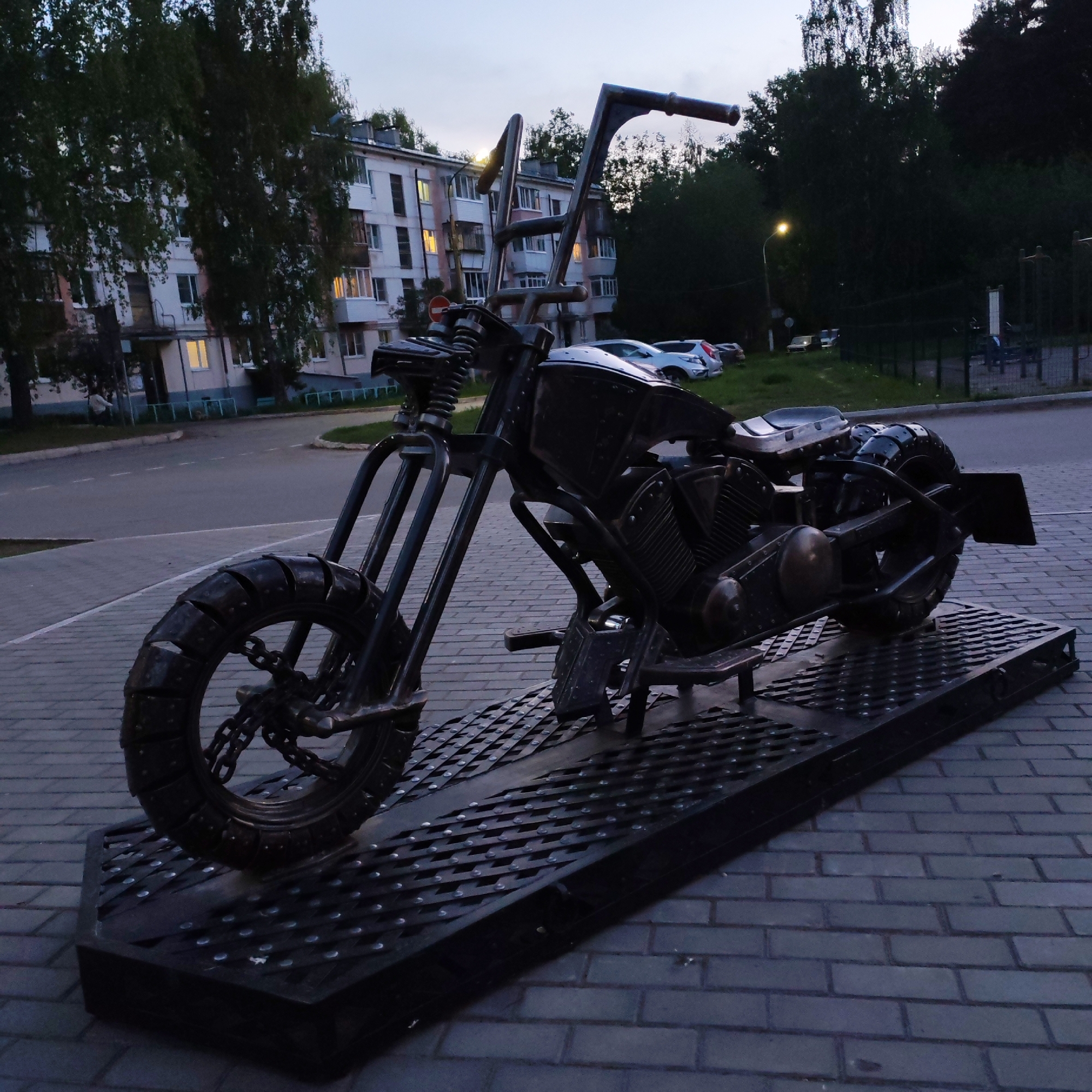 This image captures a metal sculpture of a motorcycle on display in the city of Chaikovsky, Russia. The sculpture is situated on a textured metal platform, set on a paved area. In the background, a multi-story residential building is visible, alongside trees and a street. Several cars are parked in the distance, and a lamppost illuminates the area. The time of day appears to be dusk or early evening, as the sky is dim.