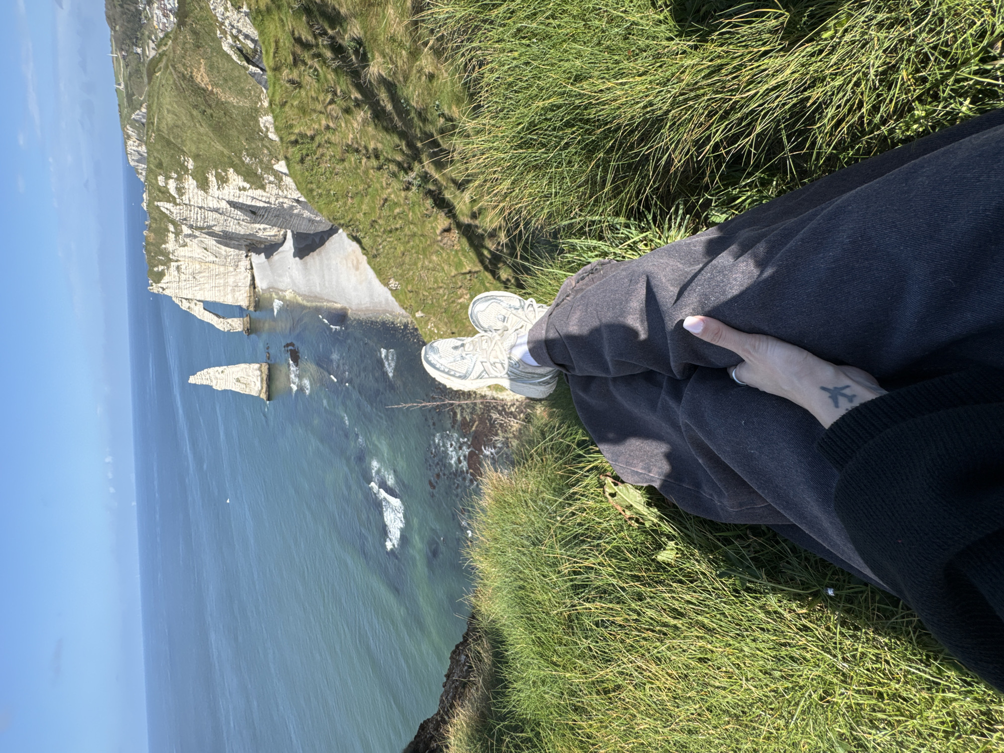 A medium, eye-level shot depicts a person seated on a grassy cliff overlooking the sea, in Étretat, France. The person is wearing grey sneakers and dark, possibly denim, trousers, with a dark sweater sleeve visible on their right. A hand rests on their leg, adorned with a tattoo of an airplane. The background reveals Étretat's distinctive chalk cliffs and sea stacks along the coastline. The ocean is a clear turquoise blue, with visible wave action. The sky is clear and blue. The immediate foreground consists of lush green grass.
