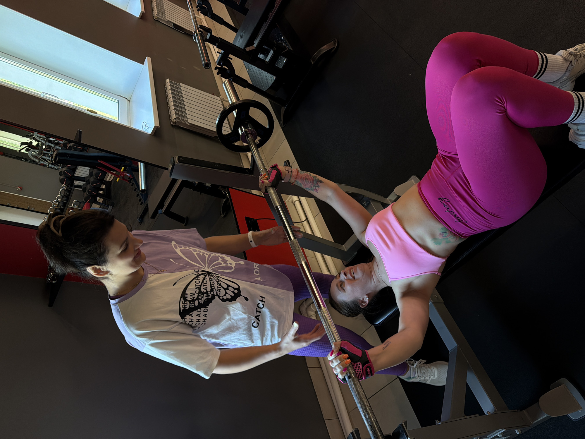 In a gym environment in Birobidzhan, Russia, two women are engaged in a weightlifting exercise. One woman, wearing a pink sports bra and bright pink leggings, is lying on a weight bench, performing a bench press with a barbell. She is wearing wrist wraps and grip gloves. The other woman, standing behind the bench, is wearing a purple and white t-shirt with a butterfly graphic and purple leggings. She appears to be spotting the woman on the bench. The gym is equipped with various weight machines and dumbbells, and natural light streams in from a large window.