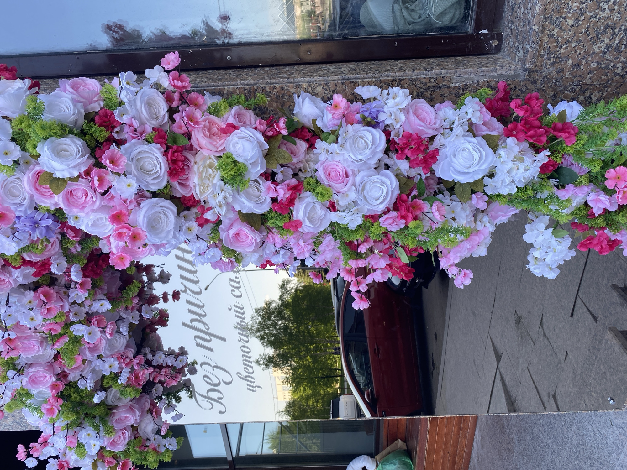 A flower arrangement composed of white, pink, and red roses, and green foliage frames the top of a sign with text in Russian. The sign is reflective, showing a distorted image of a red car, trees, and a building. A window is visible above the flower arrangement. The location is likely a business in Tyumen, Russia.