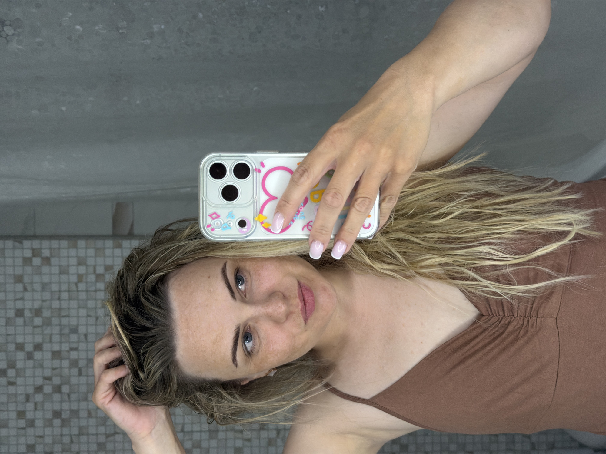 A woman is taking a selfie in what appears to be a bathroom. She is holding a smartphone with a patterned case. Her hair is blonde and wavy. She is wearing a brown top with spaghetti straps. The background includes a tiled wall and a gray surface, suggesting the bathroom setting. It is not possible to pinpoint the location to Votkinsk, Russia based on this image.