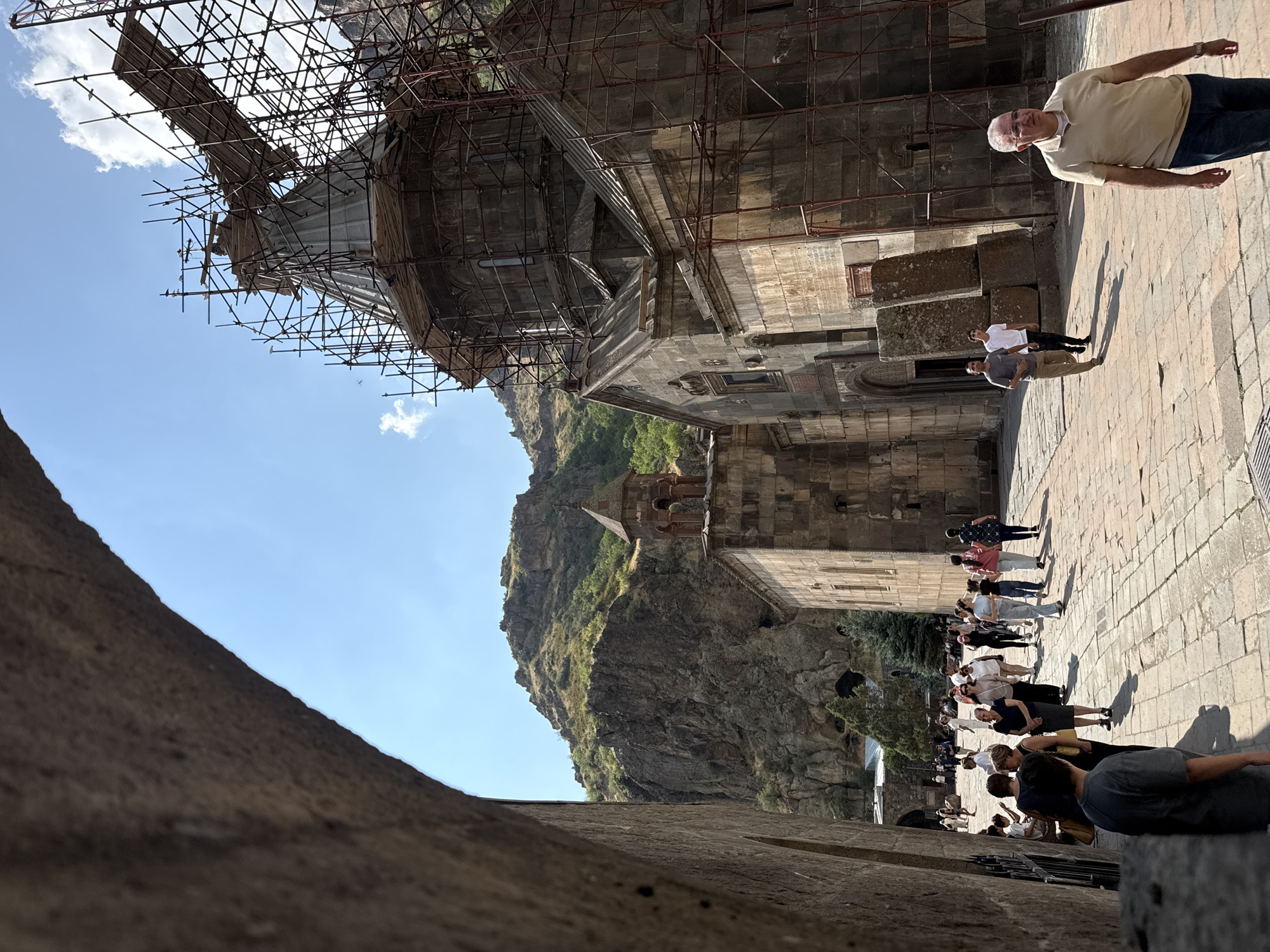 A group of tourists walk along a paved courtyard in front of ancient stone structures under a clear blue sky. In the foreground, a large arched stone structure frames the scene on the left, with the stone of its archway dominating the left side of the image. The courtyard is paved with rectangular stones.
Dominating the right side of the image is a large, multi-story stone building, possibly a church, with a domed roof. Scaffolding surrounds a significant portion of this building, obscuring its details and indicating ongoing restoration or construction work. The building's facade is made of rough-hewn stones, and it features arched windows. A smaller, bell tower-like structure with a tiered roof stands to the left of the main building, also constructed of stone.
In the background, a rugged, green mountain range rises steeply, with some trees and vegetation visible on its slopes. The mountains appear dry and rocky.
Several people are visible in the courtyard, most of whom appear to be tourists. They are walking in various directions, some in groups, others individually. One man in the mid-ground on the right wears a tan polo shirt and blue jeans. A few people are seen walking towards the large building on the right, while others are moving towards the left of the frame. A man wearing a grey t-shirt is seen in the lower left foreground, partially obscured by shadow. The lighting suggests it is daytime, with strong sunlight casting shadows. The location is the Geghard Monastery in Goght, Armenia, a UNESCO World Heritage site known for its unique architecture carved into the mountainside.