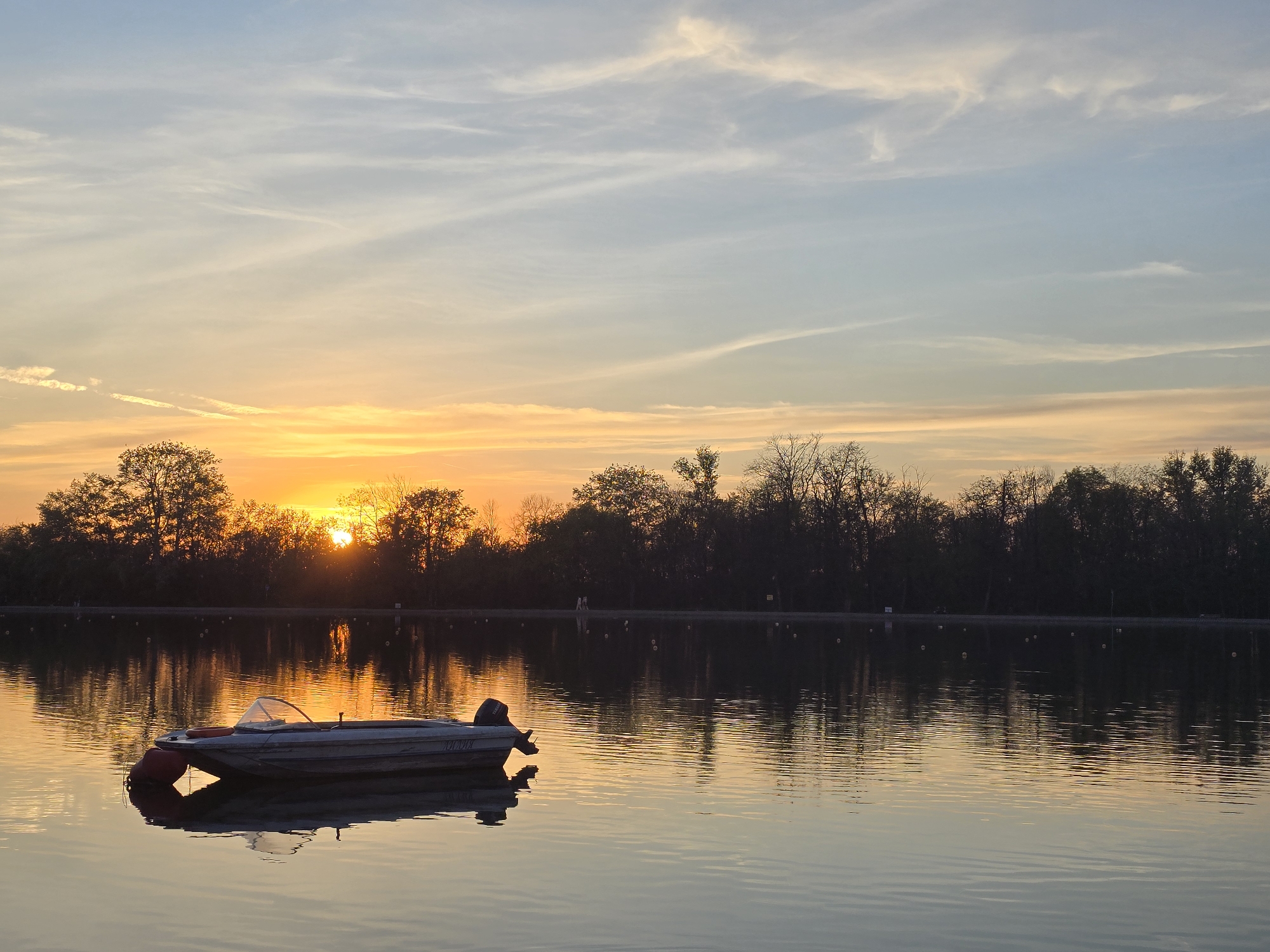 A serene sunset illuminates a tranquil lake or large pond. In the foreground, slightly to the left of the center, a small, light-colored motorboat floats calmly on the water. It features a clear windshield and a dark outboard motor at its stern, with a red buoy or fender tied to its bow. The boat's reflection is clearly visible on the still surface.
The sun, a vibrant orange, is setting just above the distant treeline on the left, casting a brilliant golden glow across the horizon and reflecting brightly on the water. The trees along the far bank are silhouetted against the radiant sky, their dark forms contrasting with the luminous background. The sky transitions from a soft blue at the top to warm oranges and yellows near the horizon, adorned with delicate, wispy clouds. The water perfectly mirrors these colors and the silhouettes of the trees and sky, creating a harmonious and peaceful scene.
Small white or yellow buoys are scattered across the water near the distant bank, possibly marking a perimeter. Faint human figures can be discerned walking along the far bank. The time of day is clearly sunset, and the weather appears calm and clear. The overall impression is one of quiet beauty and repose. On the side of the boat, the Cyrillic word "ЛАДЬЯ" is visible.