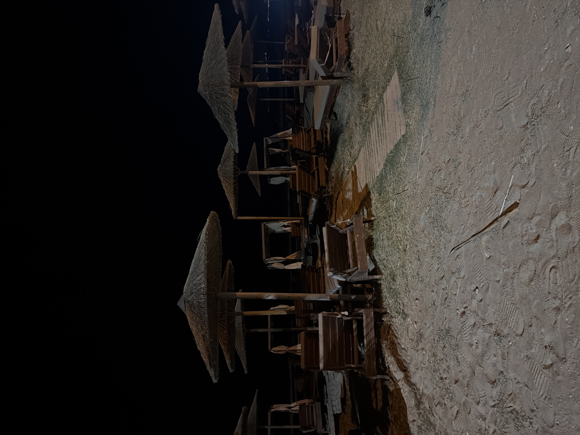 At night on a sandy beach in Georgioupoli, Greece, several empty sun loungers are arranged under thatched umbrellas. The beach is lit by artificial lights, casting shadows and highlighting the texture of the sand and dry grass. In the background, a few distant lights suggest the presence of a town or village. A wooden walkway is visible leading through the scene.