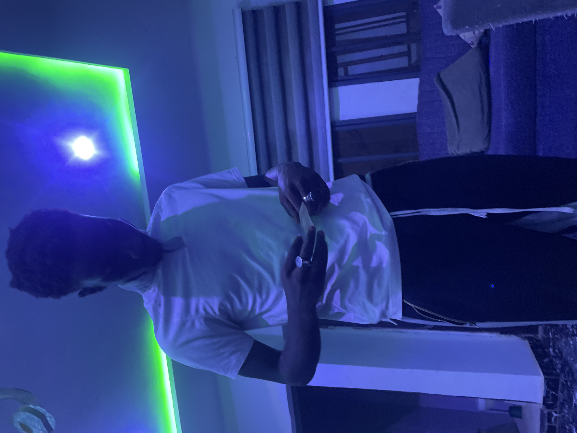 The image, rotated 90 degrees counter-clockwise, displays an individual standing in an indoor setting, heavily illuminated by artificial light. The subject, likely male, is wearing a white short-sleeved t-shirt and dark trousers. Their face is obscured by shadow and angle. They are holding a small, light-colored object, possibly a phone or remote control, in both hands near their waist. Several rings are visible on their fingers.
The setting is a room characterized by dynamic lighting, predominantly in shades of blue and green. A prominent neon green light strip runs along what appears to be a wall or ceiling fixture on the left, casting a vibrant glow. A bright, white light source, resembling a starburst, is also visible within this green illuminated area. The remainder of the room is bathed in a cool blue light, suggesting extensive use of LED or neon lighting. In the background, elements such as vertical blinds or drapes over a window and parts of furniture (possibly a bed or couch, and a cabinet or shelf) are dimly visible. The dark ambient conditions imply the image was taken at night or in a purposefully darkened room to highlight the artificial lights. There is no visible text.