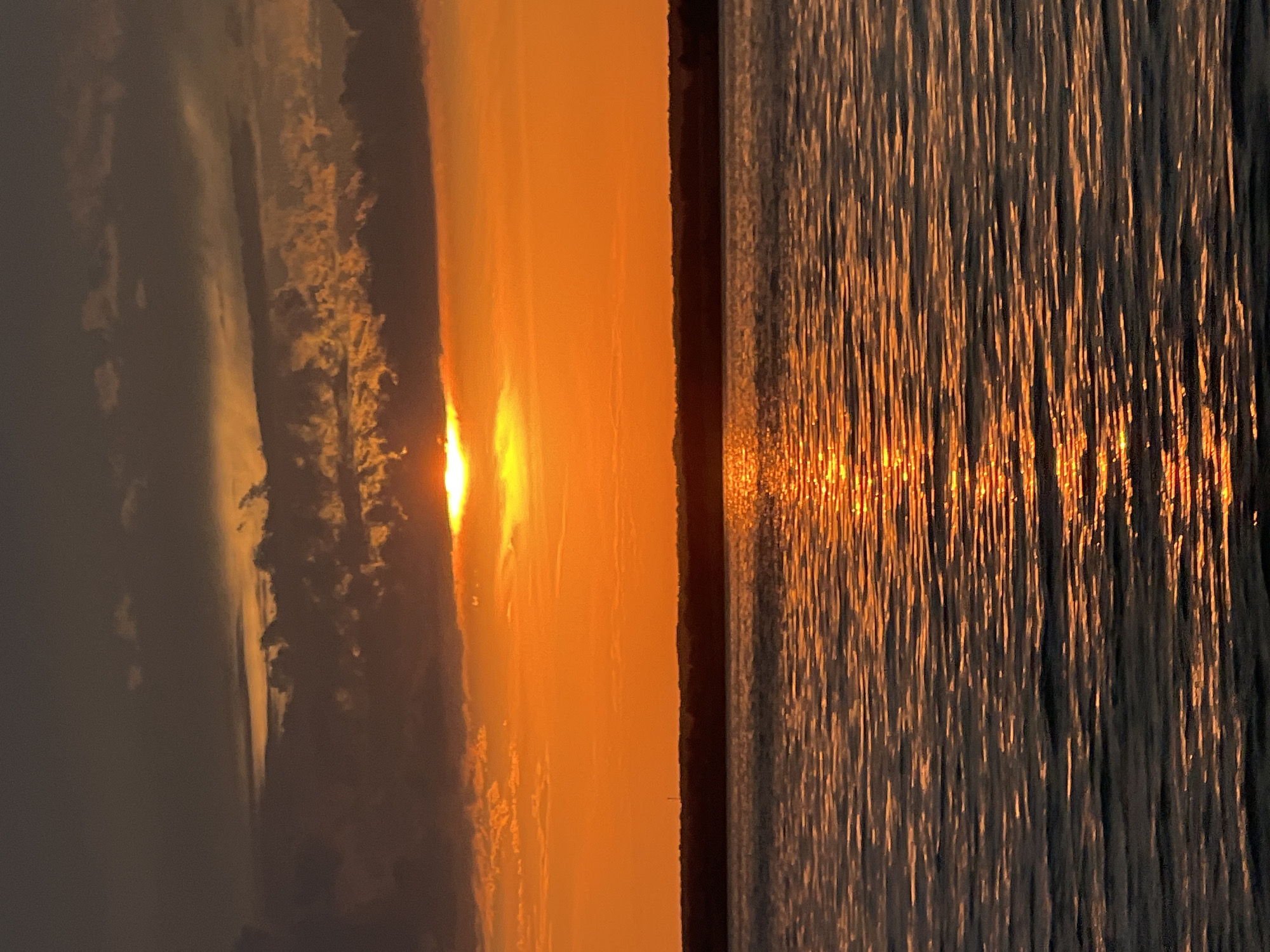 The image depicts a natural landscape at sunset over a body of water, likely the Black Sea, near Beregovoe, Russia. The sky is dominated by a vibrant orange glow from the setting sun, which is partially obscured by dark, textured clouds. A sliver of the sun is visible peeking through the cloud cover, casting a brilliant reflection on the water's surface. The water itself is rippled, with the sunset's light creating shimmering streaks of orange and gold across its dark, calm expanse. A dark, silhouetted line of trees or landmass forms the horizon between the sky and the water. There are no people or man-made objects visible in the scene.