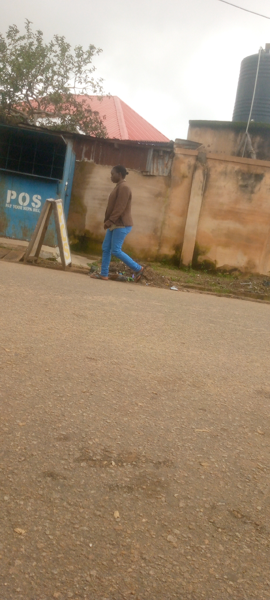 A young woman walks along a paved road in an urban setting. She is wearing blue jeans and a brown jacket, and her hair is pulled back. To her left, a blue building with the word "POS" painted on it is visible, along with an A-frame sign. Behind the building, a red-roofed structure and a large water tank can be seen. The road surface is rough and discolored, with some debris scattered along the side. The sky is overcast.