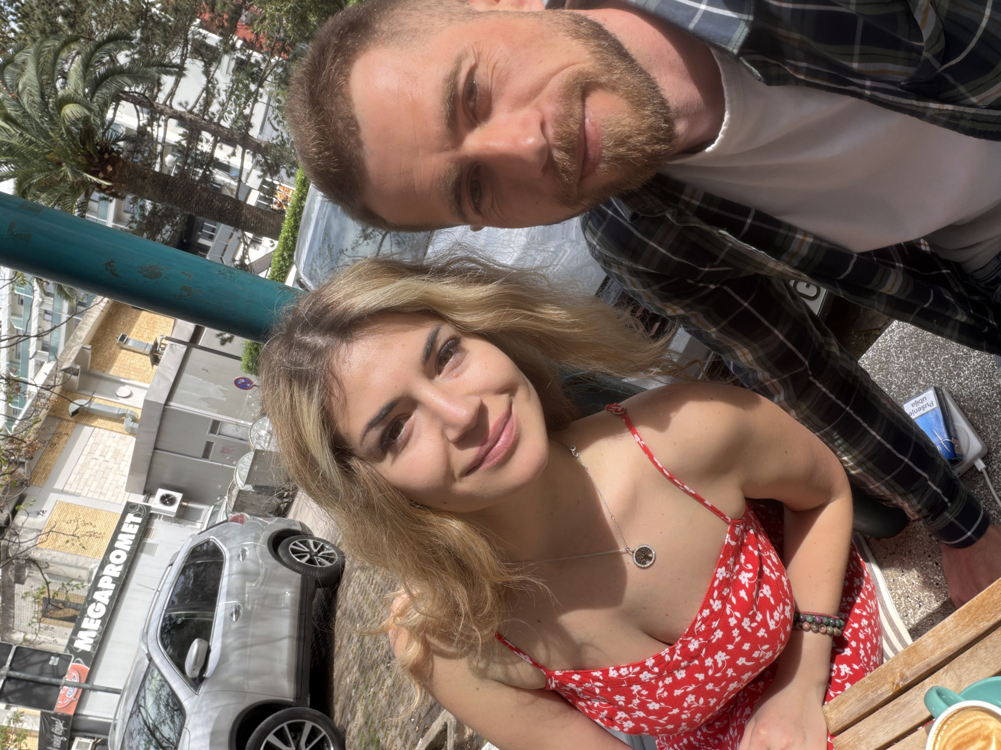 A man and a woman are photographed in an outdoor setting, likely a cafe in the city of Budva, Montenegro, as "MEGAPROMET" is visible on a building in the background. The woman is in the foreground wearing a red floral dress and a silver necklace. The man is behind her, wearing a white t-shirt and a plaid shirt. A palm tree and other greenery are visible in the background. A blue object, possibly a metal pole, is also visible in the background. A car, likely parked on the street, and the corner of a wooden table with a cup of coffee are also visible.
