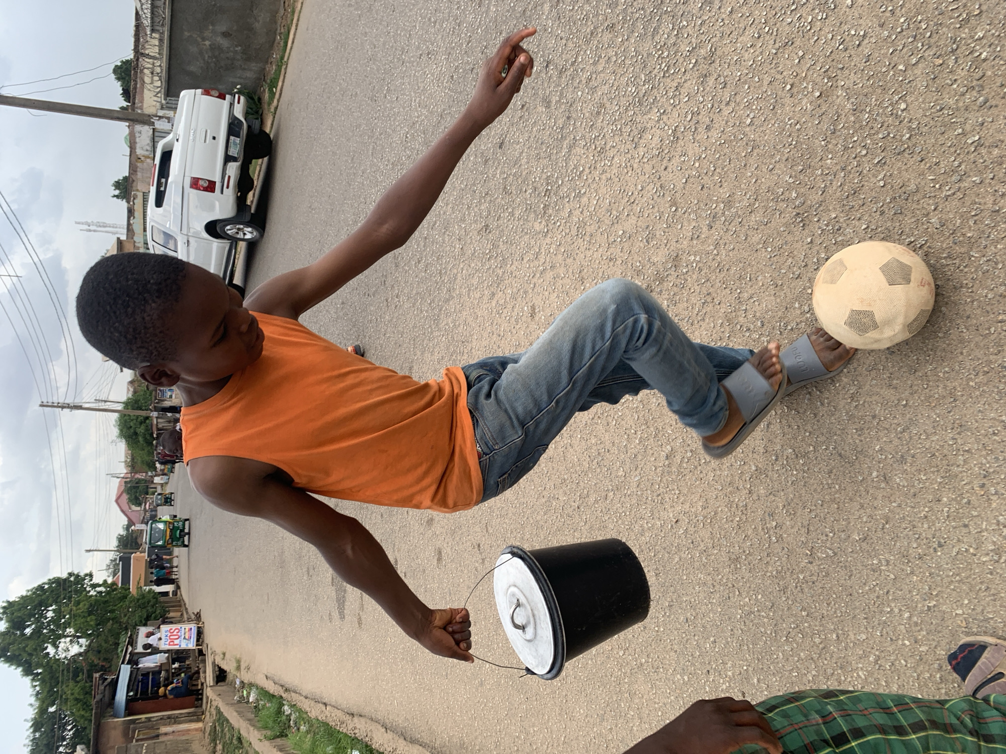 A young man is walking on a paved street in Jos, Nigeria. He is wearing an orange sleeveless shirt, blue jeans, and grey sandals. He is carrying a black bucket with a silver lid in his left hand and gesturing with his right hand. A soccer ball is near his feet. A white pickup truck is parked on the side of the street. In the background are buildings, trees, and traffic including motorized tricycles. Another person is partially visible in the lower right corner.