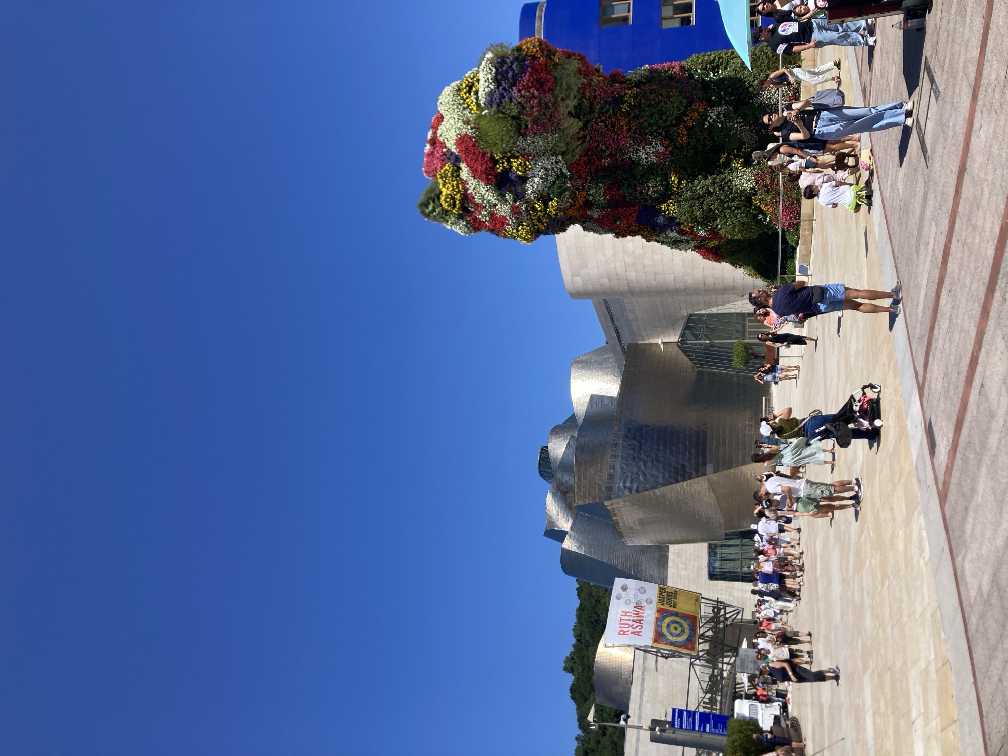 The image depicts the exterior of the Guggenheim Museum Bilbao, a renowned architectural landmark in Bilbao, Spain. In the foreground, a large, vibrant floral sculpture, likely a temporary installation, dominates the scene. The sculpture is composed of a multitude of colorful flowers and greenery, forming an abstract, organic shape. A diverse crowd of people is spread across the plaza in front of the museum, engaged in various activities. Some are walking, others are standing and conversing, and a few appear to be taking photographs.
The Guggenheim Museum itself is characterized by its striking, undulating metallic facade, which reflects the bright sunlight. Behind the main museum building, a smaller, cylindrical blue structure is visible. The sky is a clear, deep blue, indicating fair weather. Several banners and signs are displayed on the museum's exterior, one of which reads "RUTH ASAWA" and another "JASPER JOHNS." The plaza is paved with light-colored stone tiles, and benches are scattered throughout the area. The overall atmosphere suggests a busy day at a popular tourist attraction.