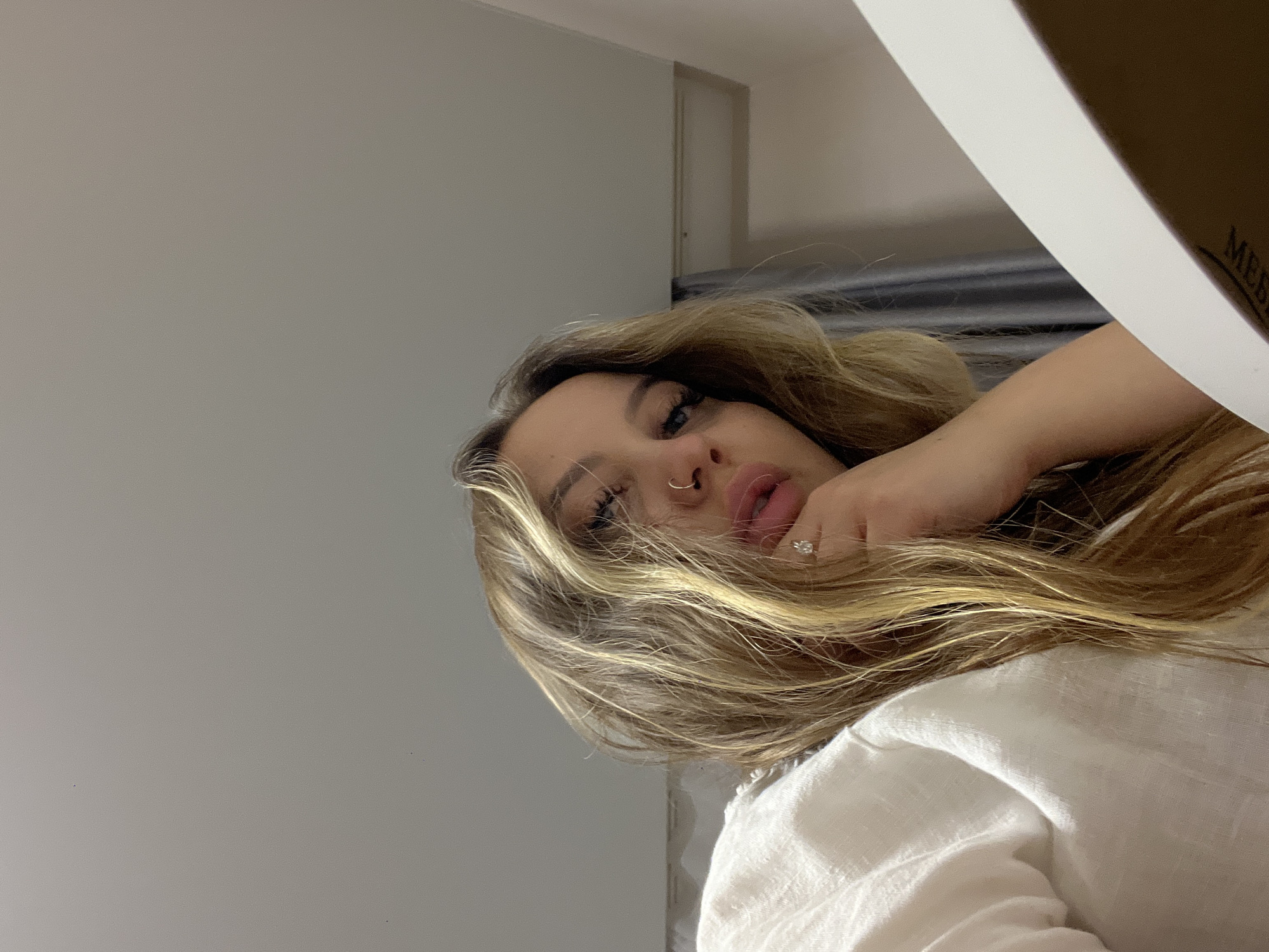 A young woman with long blonde hair and a nose ring is photographed from a low angle. She is wearing a white shirt or top. Her hand is near her face, and she is wearing a ring. The background is a neutral gray wall, possibly in a room. The presence of a bed or bedding suggests the setting is a bedroom. The image appears to be taken indoors with artificial lighting. There is no information available to determine the location is in Pritsepilovskaya, Russia.