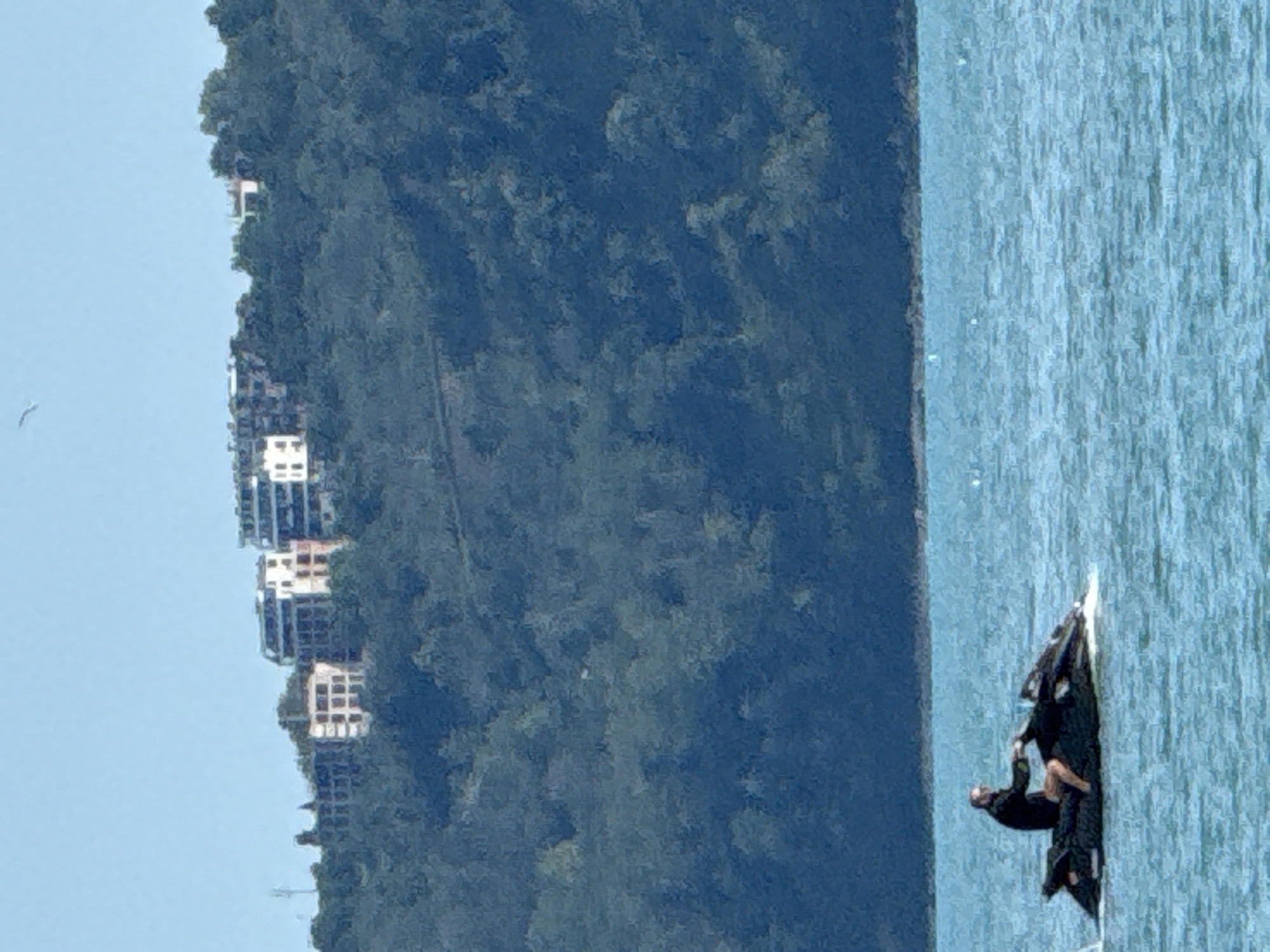 Location: Unknown, Unknown
The image captures a daytime scene featuring a body of water, a forested hillside, and buildings under a clear sky.
In the foreground, a single male individual is operating a black jet ski on a body of water with a turquoise hue and a textured, rippled surface. The man is positioned centrally on the jet ski, wearing a dark, long-sleeved top, dark shorts, and dark eye protection (sunglasses or goggles). His hands are on the handlebars, and the jet ski is generating a small white wake behind it, indicating movement.
The midground consists of a densely vegetated hillside, characterized by dark green foliage, suggesting a forest. The hill rises steeply from the water's edge.
On the crest of the hillside in the background, a cluster of several multi-story residential or commercial structures is visible. The buildings primarily feature light-toned facades (white or cream), often contrasted by dark window or balcony elements. One prominent structure on the mid-left has a distinct grid-like pattern and appears darker, possibly brownish or grey. To the far left, a slender, dark vertical structure (possibly an antenna or utility pole) protrudes slightly above the tree line. Above the buildings, the sky is clear, pale blue, and devoid of clouds. A single white bird is discernible, soaring in the upper-mid portion of the sky.