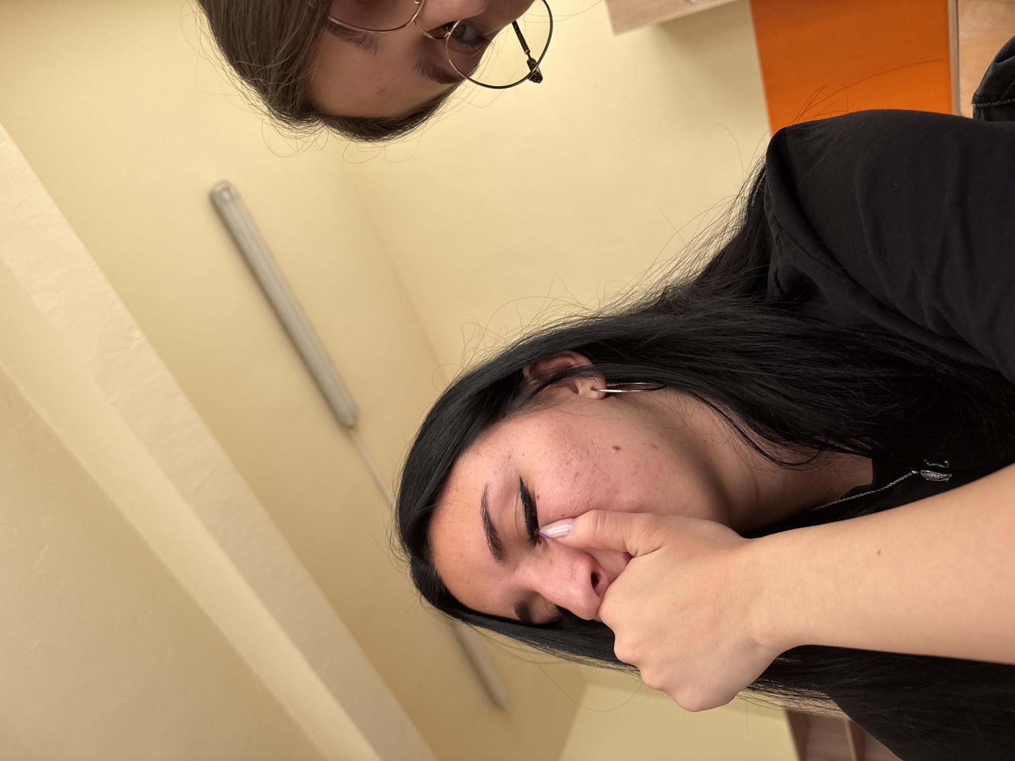 Two individuals are captured from an elevated, angled perspective. One person's face, partially visible, is framed by dark hair and round eyeglasses. The other individual, with long dark hair and an earring, appears to be touching their face with their hand. The background consists of a light yellow wall and an orange-colored surface. There is no information available to determine if the location of the photo is in Sofia, Bulgaria, or any other city.